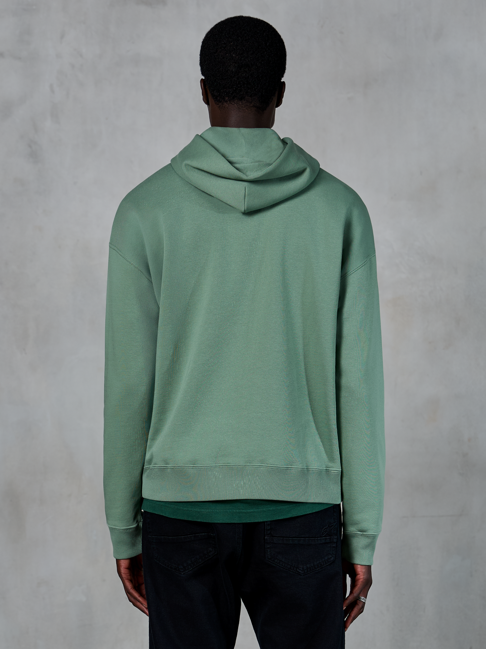 Alpinestars Essentials Ageless Hoodie, avslappnade sportkläder, kaktusgrön, sedd bakifrån på modell, plaggstenstvättad finish, matchad med svarta byxor, minimalistisk livsstilsdesign