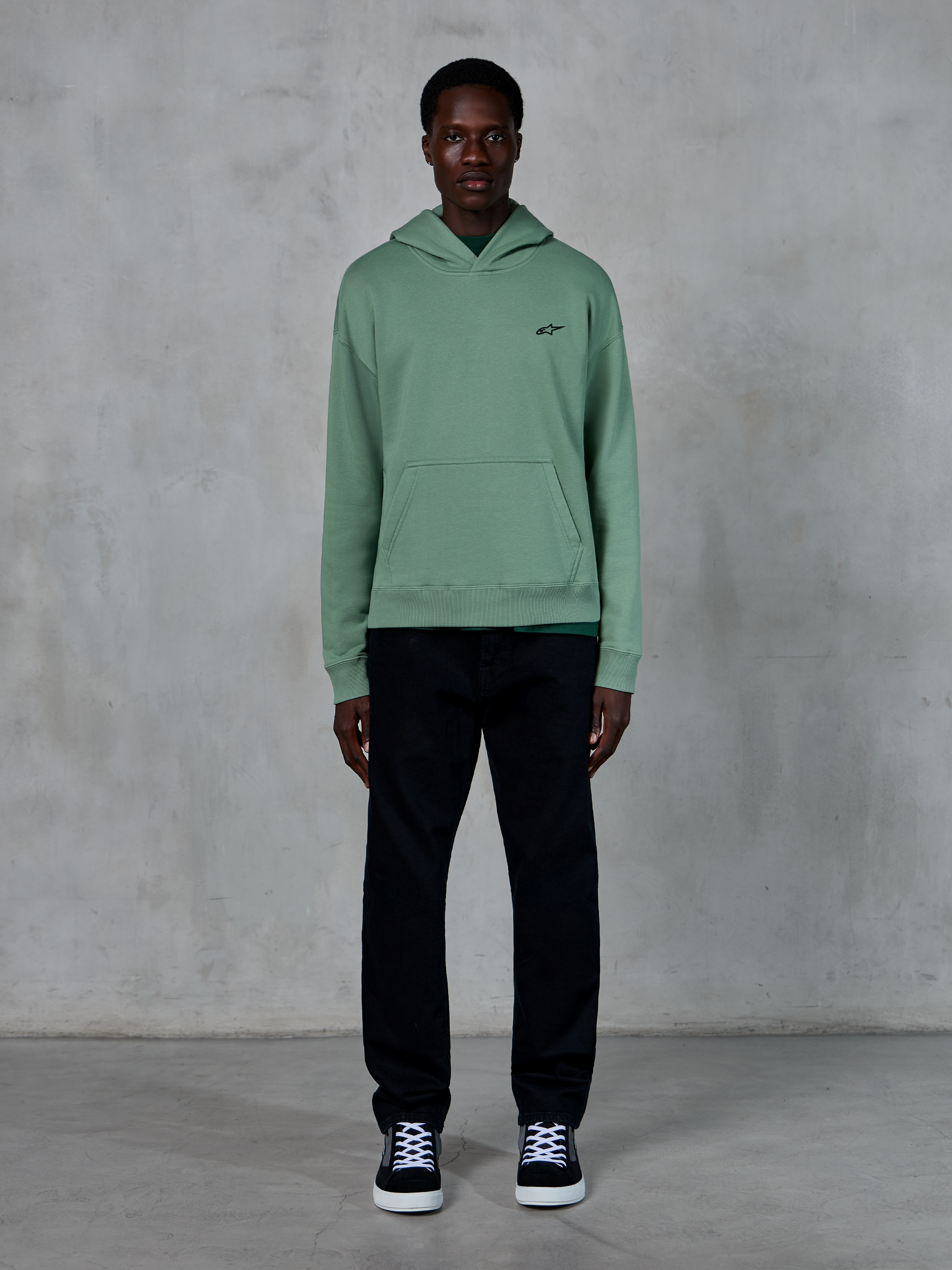 Alpinestars Essentials Ageless Hoodie, kaktusgrön, liten svart logotyp på bröstet, känguruficka, avslappnad lifestyle-streetwear som bärs med svarta byxor och svartvita sneakers mot en texturerad vägg.