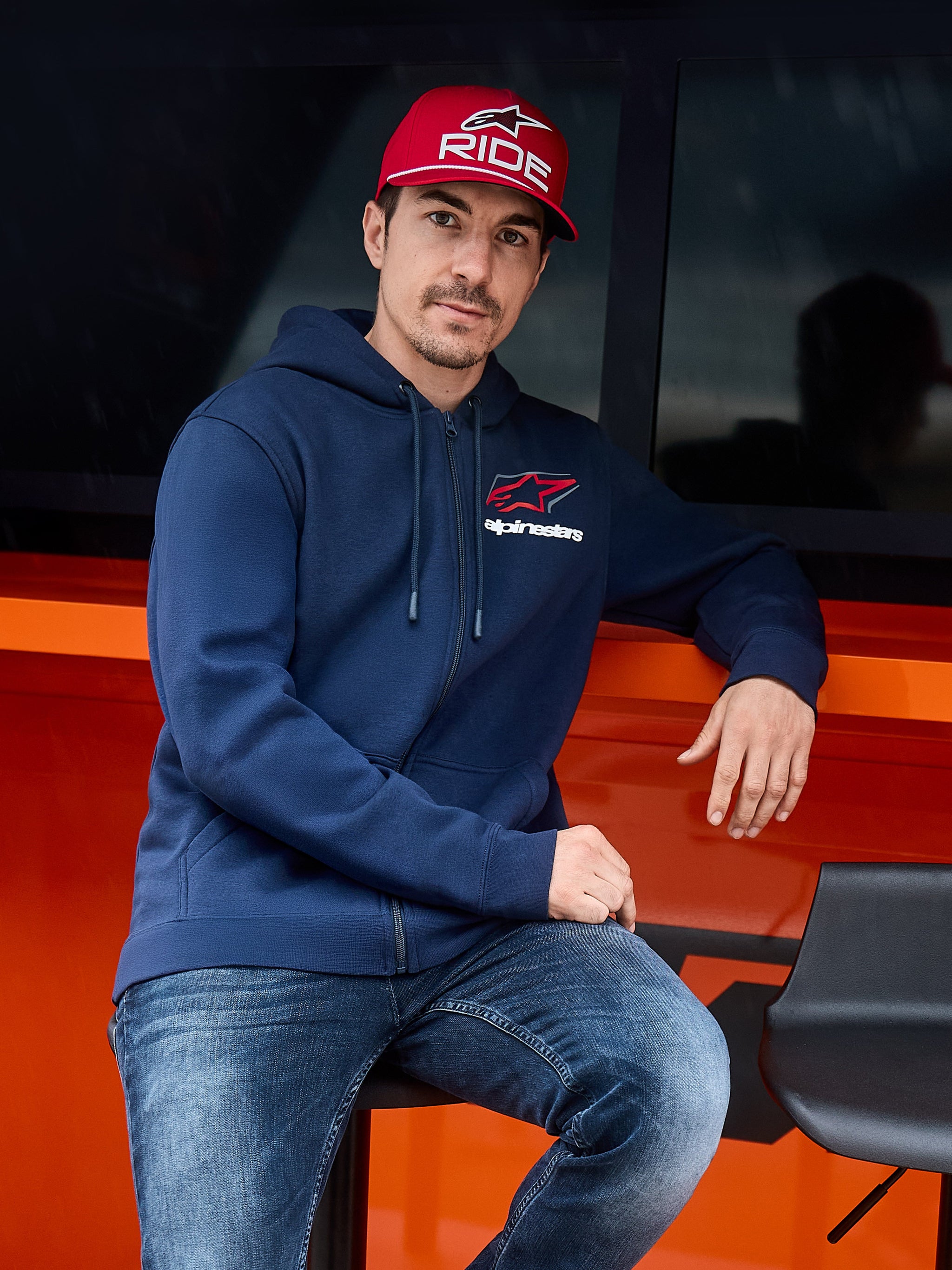 Alpinestars Formulation Huvtröja, marinblå, huvtröja med dragkedja framtill och Alpinestars-logotyp på bröstet, bärs med en röd keps och jeans, avslappnad motorsport-livsstil streetwear