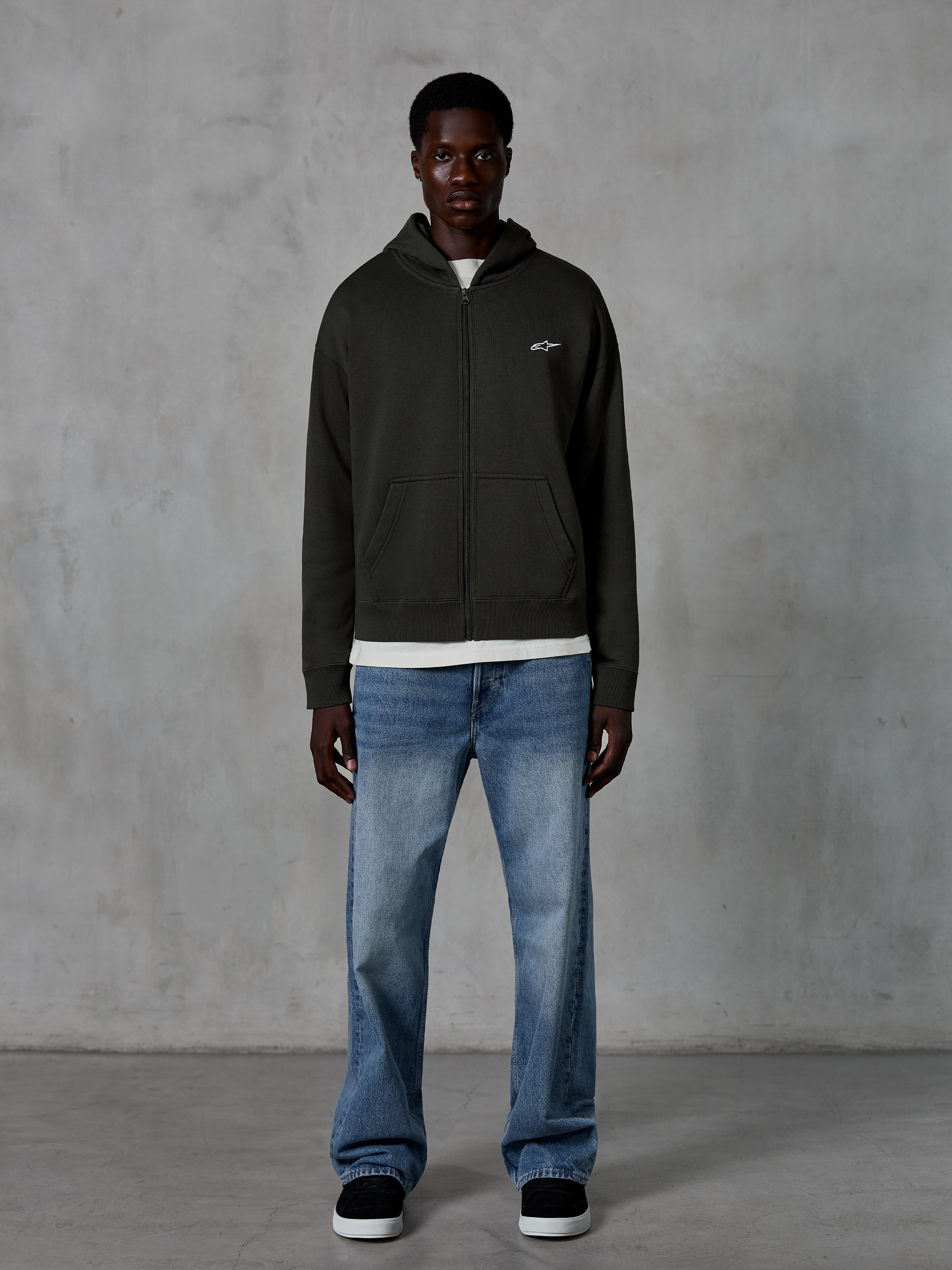 Alpinestars Essentials Ageless Zip Hoodie, huvtröja med hel dragkedja, Off Black, med en liten vit Alpinestars-logotyp på bröstet, bärs av en modell med en vit t-shirt och blå jeans, avslappnad passform, casual motorsport-streetwear