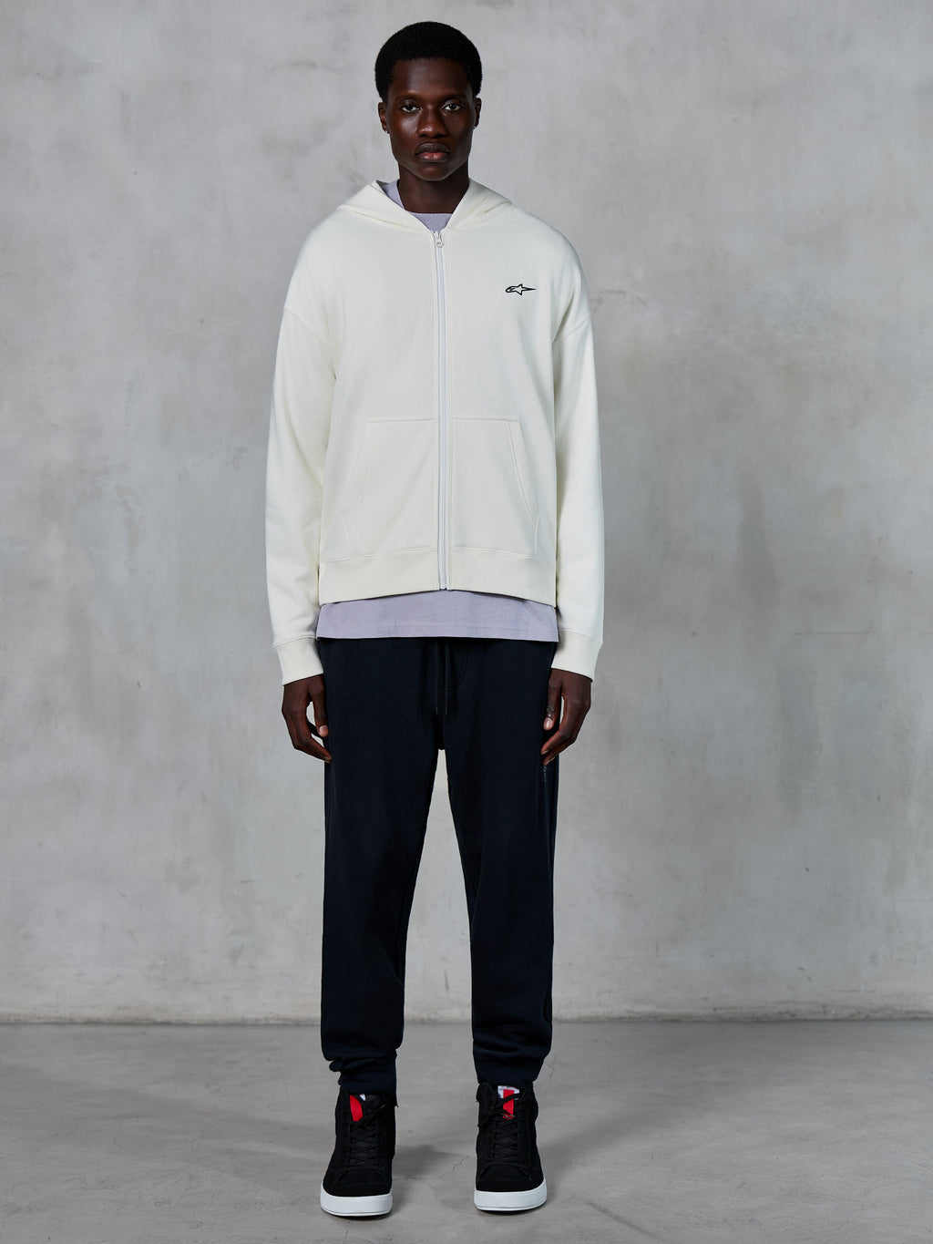 Alpinestars Essentials Ageless Zip Hoodie, benvit huvtröja med dragkedja, avslappnad passform med en liten svart logotyp på bröstet, bärs med svarta mjukisbyxor och sneakers, avslappnat motorsportinspirerat streetwear.