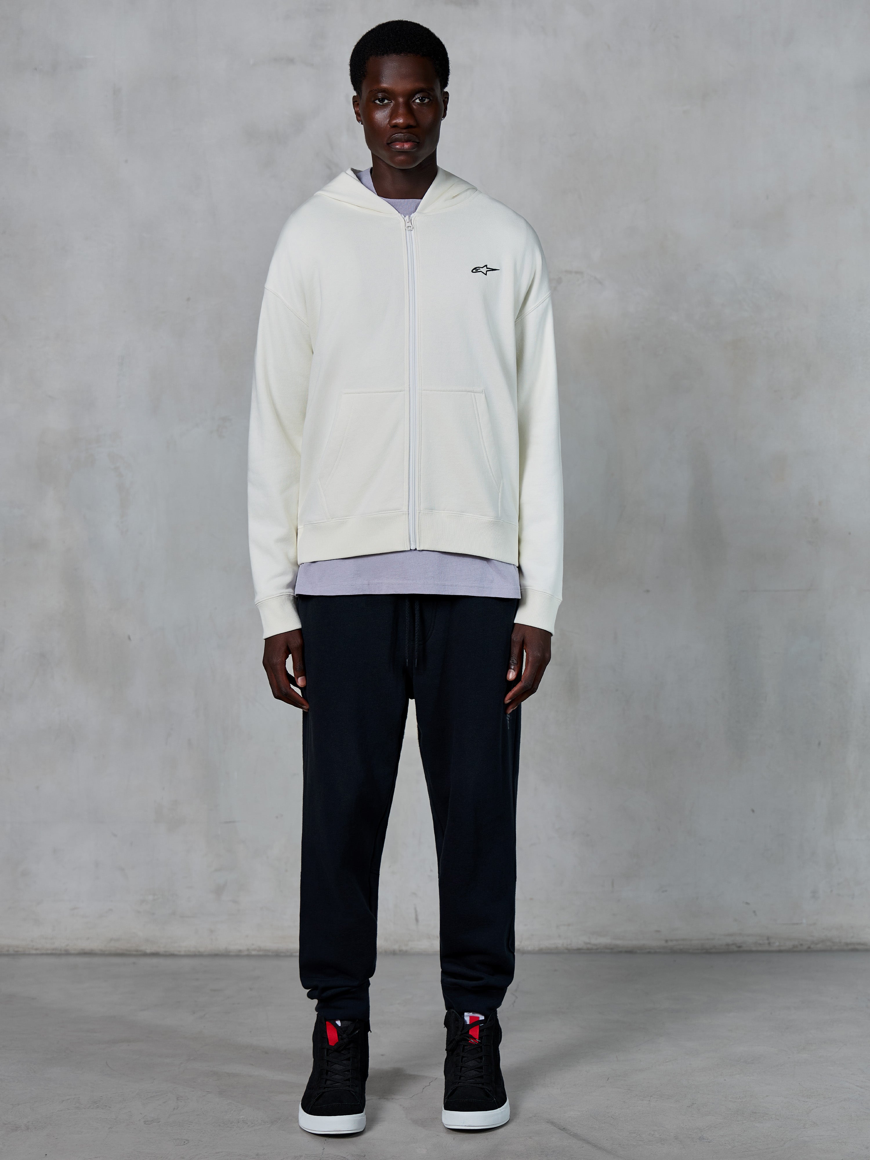 Alpinestars Essentials Ageless Zip Hoodie, benvit huvtröja med dragkedja, avslappnad passform med en liten svart logotyp på bröstet, bärs med svarta mjukisbyxor och sneakers, avslappnat motorsportinspirerat streetwear.