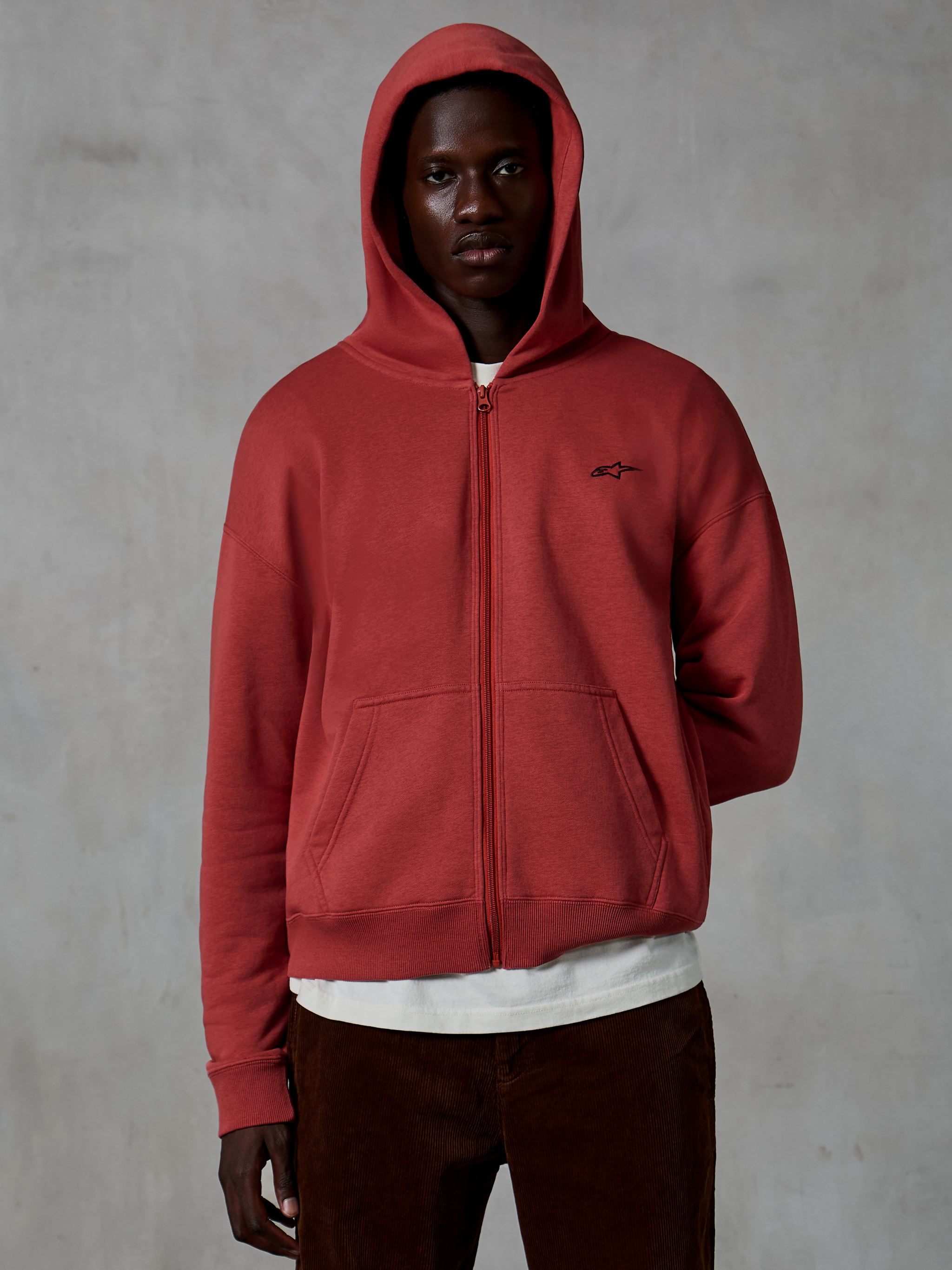 Alpinestars Essentials Ageless Zip Hoodie, avslappnade sportkläder i tegelrött, design med hel dragkedja och uppfälld huva, med en liten svart logotyp på bröstet, bärs av en modell med en vit t-shirt och bruna manchesterbyxor mot en grå bakgrund.
