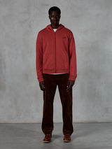 Alpinestars Essentials Ageless Zip Hoodie, casual streetwear, tegelröd med liten svart stjärnlogotyp, dragkedja framtill, bärs av en modell med bruna manchesterbyxor och kängor, premium motorsport-inspirerad design