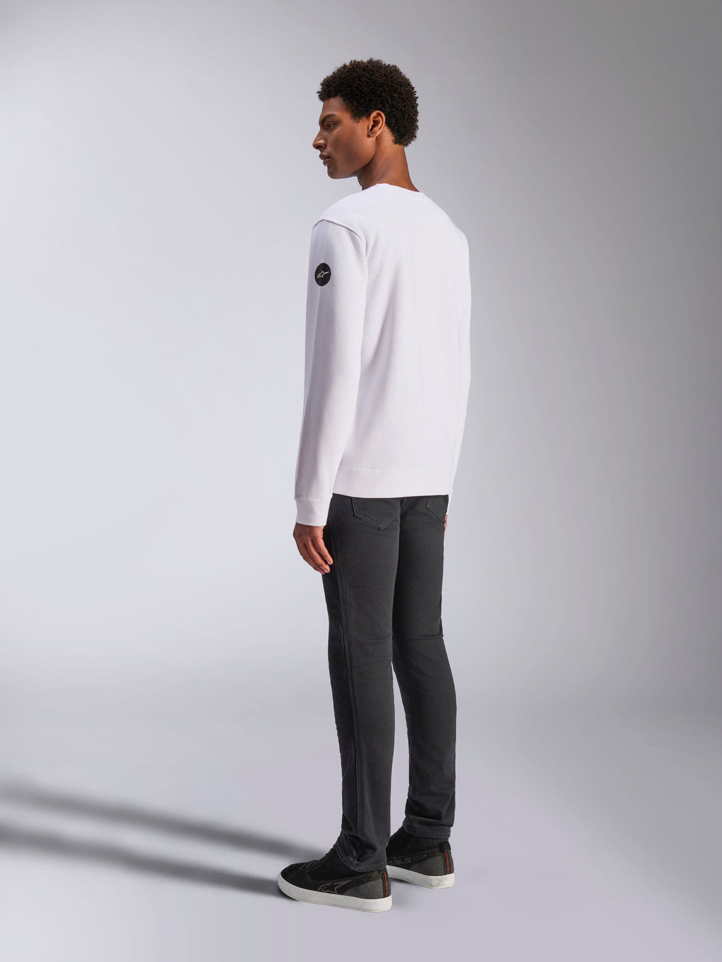 Alpinestars Bettern Crew, vit sweatshirt med rund halsringning, bak- och sidovy, svart rund logotyp på ärmen, bärs med mörkgrå byxor och svarta sneakers, avslappnad motorsport-streetwear
