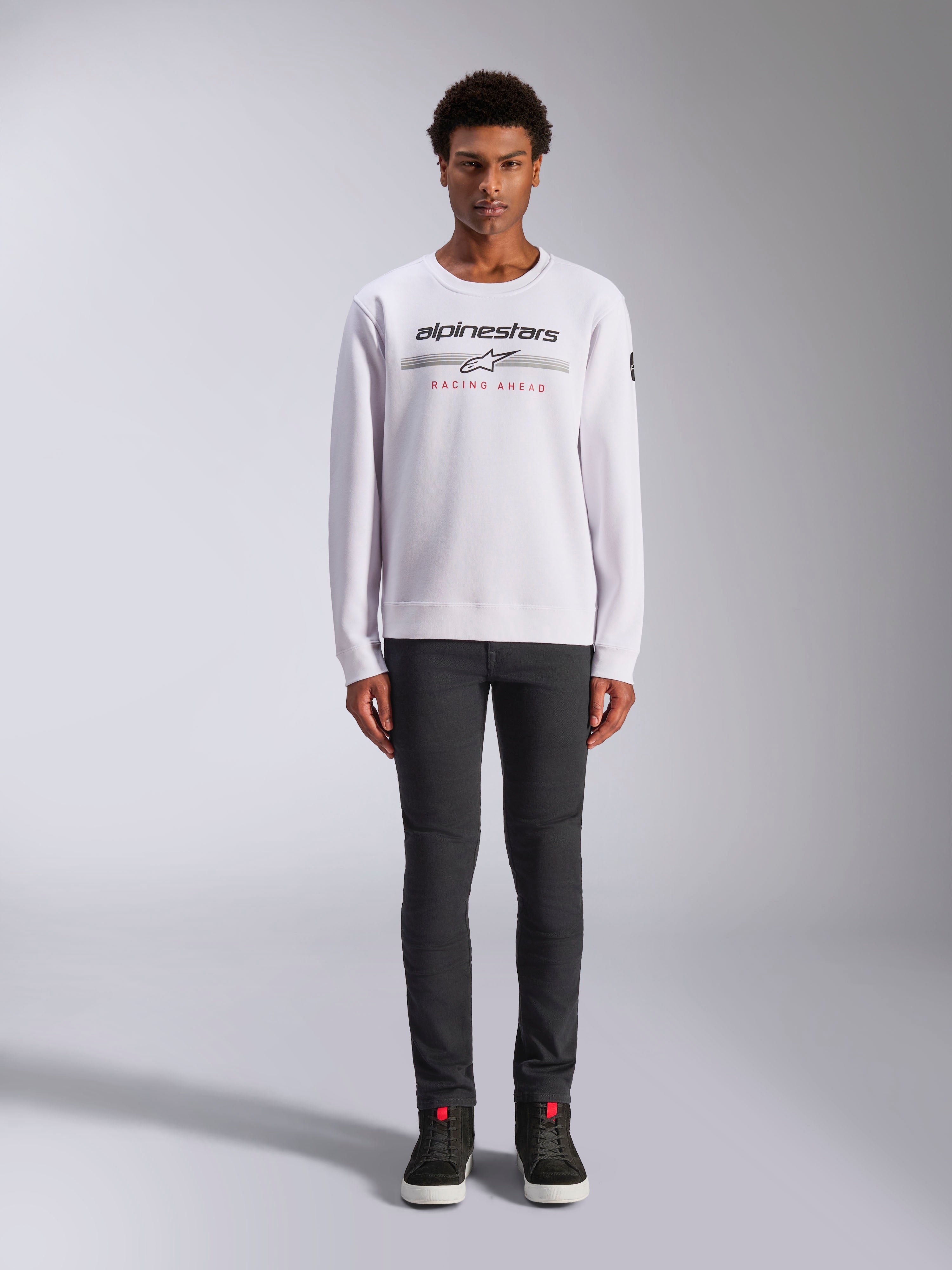 Alpinestars Bettern Crew, vit sweatshirt med rund hals, bärs av en manlig modell med mörkgrå byxor och svarta high-top sneakers, med Alpinestars-logotyp och röd 'Racing Ahead'-text, avslappnad motorsportinspirerad streetwear