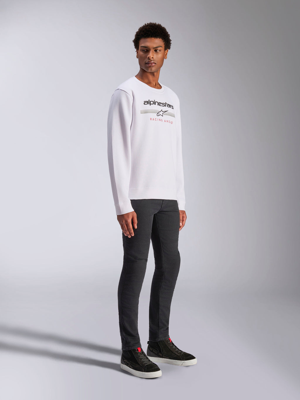 Alpinestars Bettern Crew, vit sweatshirt, med svart Alpinestars-logotyp och röd 'Racing Ahead'-text, casual motorsport-streetwear, buren av en modell med mörka byxor och svarta sneakers
