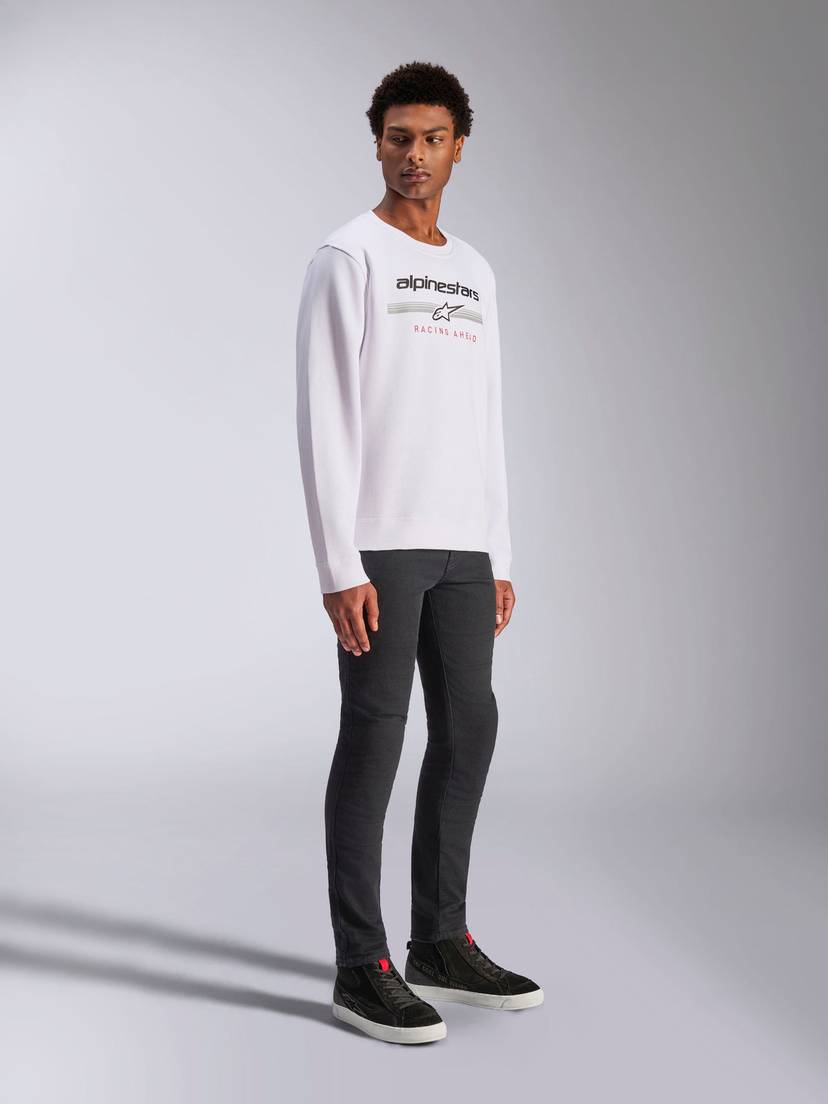 Alpinestars Bettern Crew, vit sweatshirt, med svart Alpinestars-logotyp och röd 'Racing Ahead'-text, casual motorsport-streetwear, buren av en modell med mörka byxor och svarta sneakers