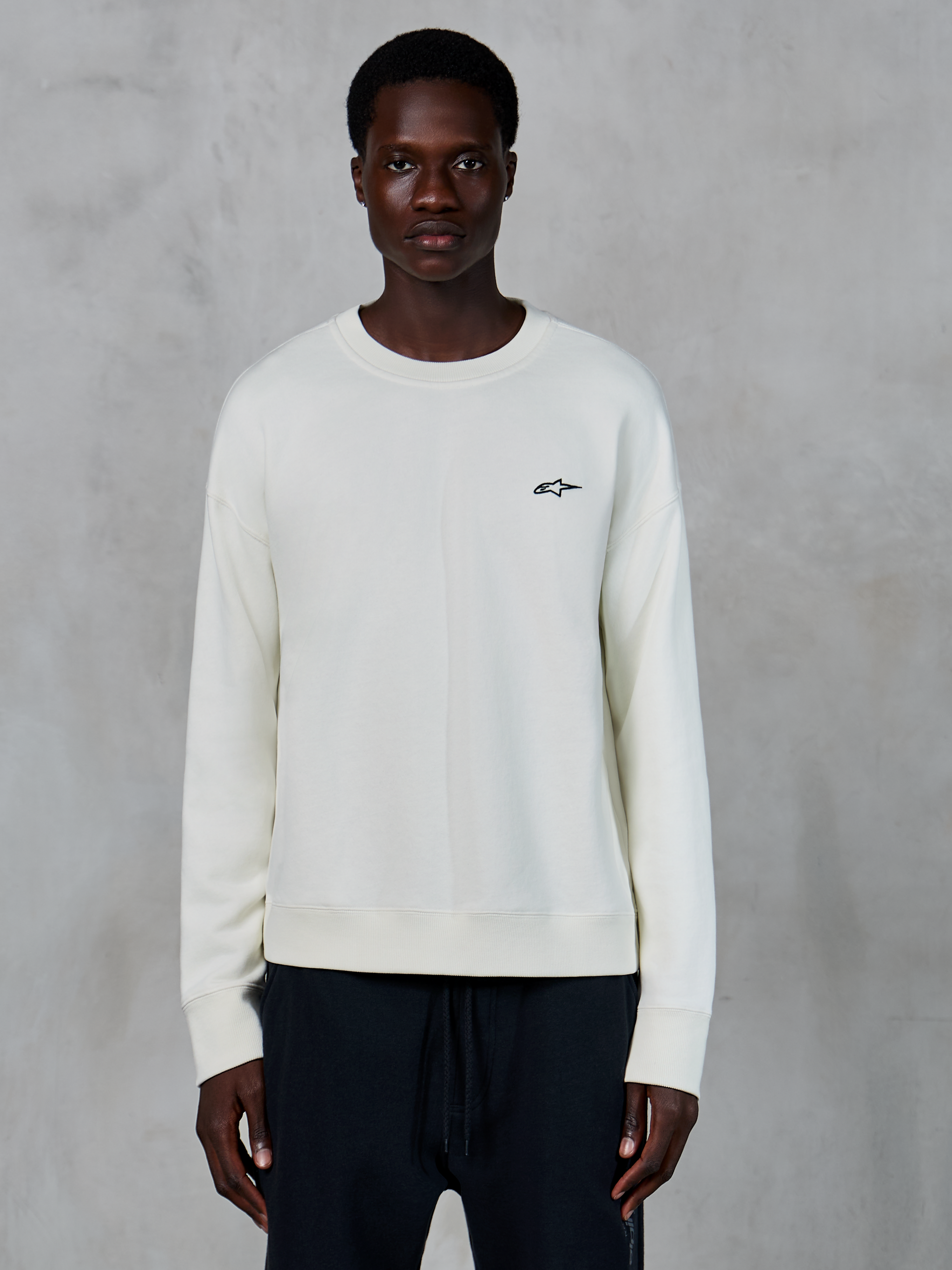 Alpinestars Essentials Ageless Crew, Off White crewneck-sweatshirt, liten svart Astars-logotyp på vänster bröst, casual livsstilssportkläder, premium fleecestruktur, bärs av modell med svarta joggers