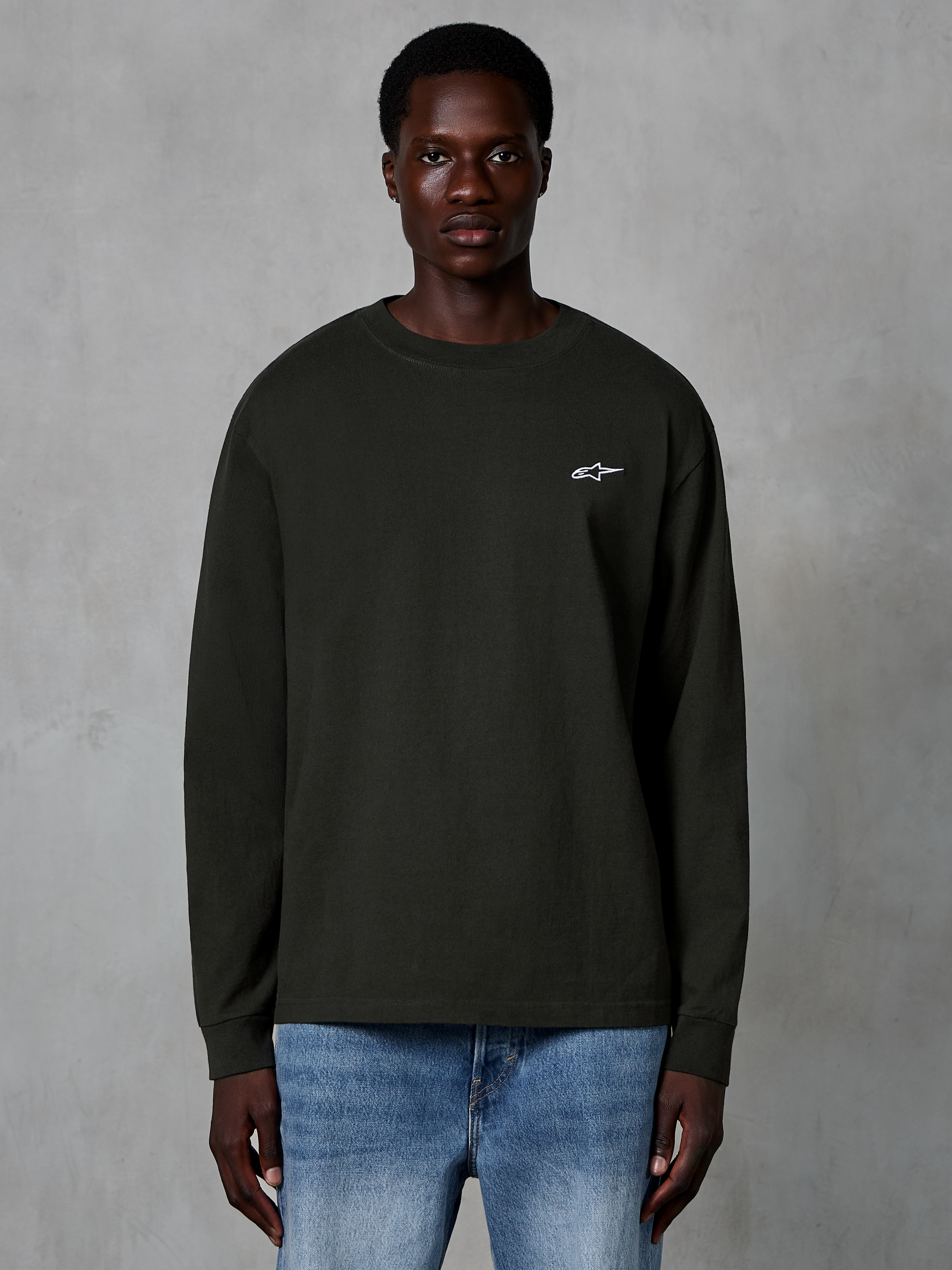 Alpinestars Essentials Ageless LS Tee, långärmad t-shirt, Off Black, med en liten vit Alpinestars-logotyp på bröstet, bärs av en modell med blå jeans, vardaglig streetwear i ekologisk bomull