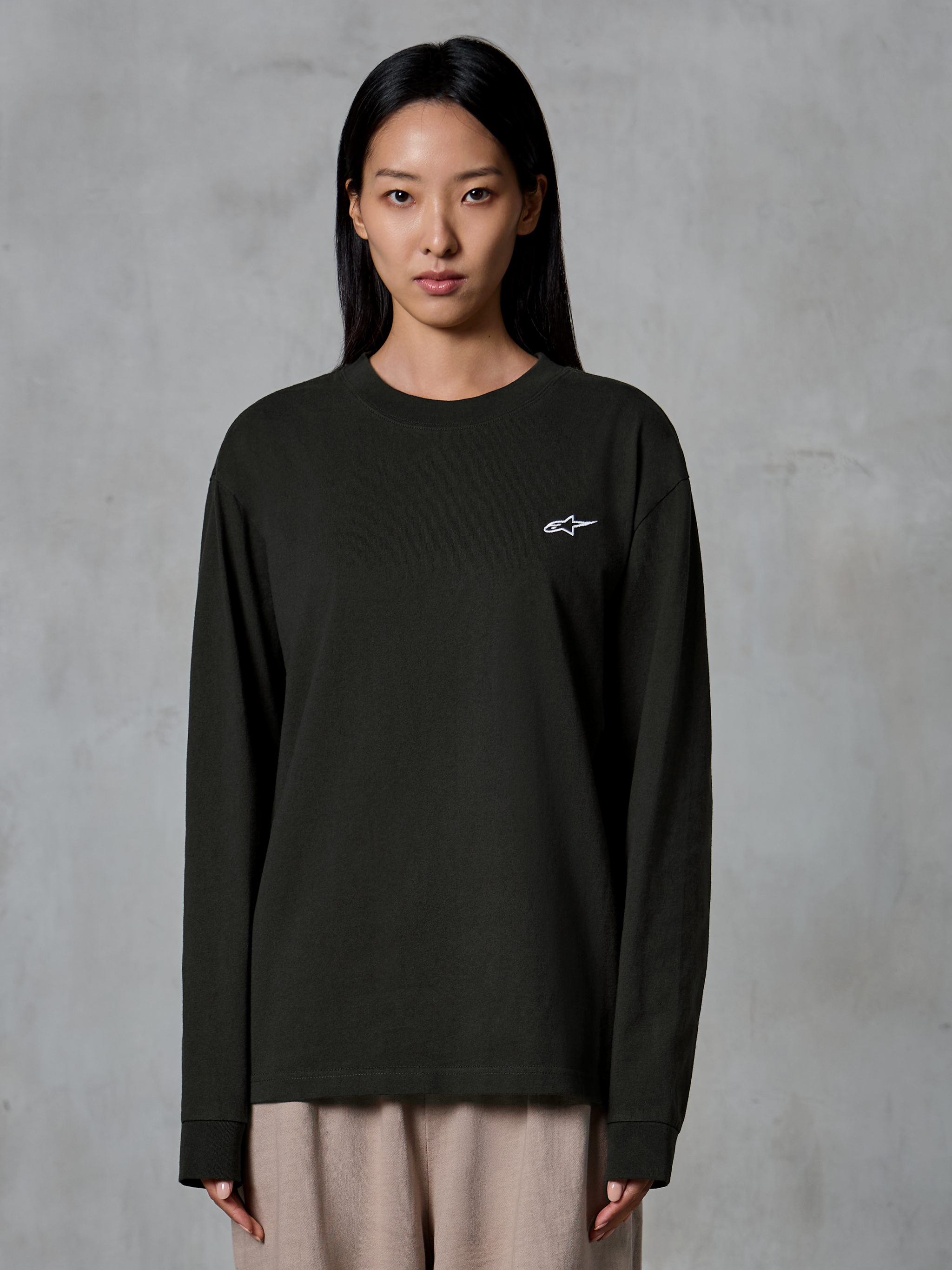 Modell som bär Alpinestars Essentials Ageless LS Tee, långärmad t-shirt, Off Black, med en liten vit Alpinestars-logotyp på bröstet, kombinerad med beige byxor, avslappnad lifestyle-streetwear