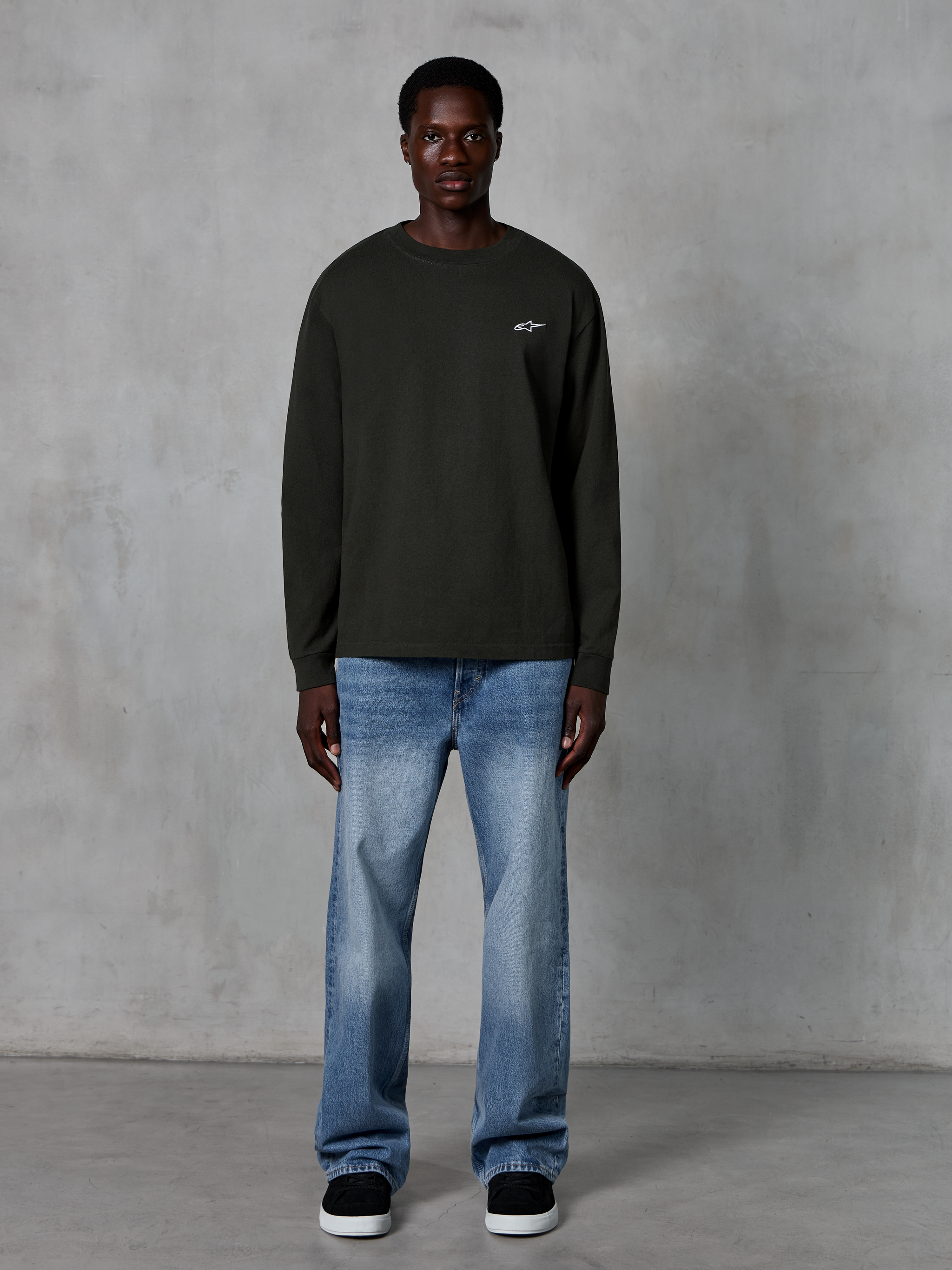 Alpinestars Essentials Ageless LS Tee, långärmad t-shirt, Off Black, med en liten vit stjärnlogotyp på bröstet, avslappnad passform, casual motorsport-streetwear, stylad med ljusblå jeans