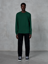 Alpinestars Essentials Ageless LS Tee, långärmad t-shirt, tallgrön, med en liten svart logotyp på bröstet, bärs av en modell med svarta byxor och sneakers, avslappnad streetwear-stil