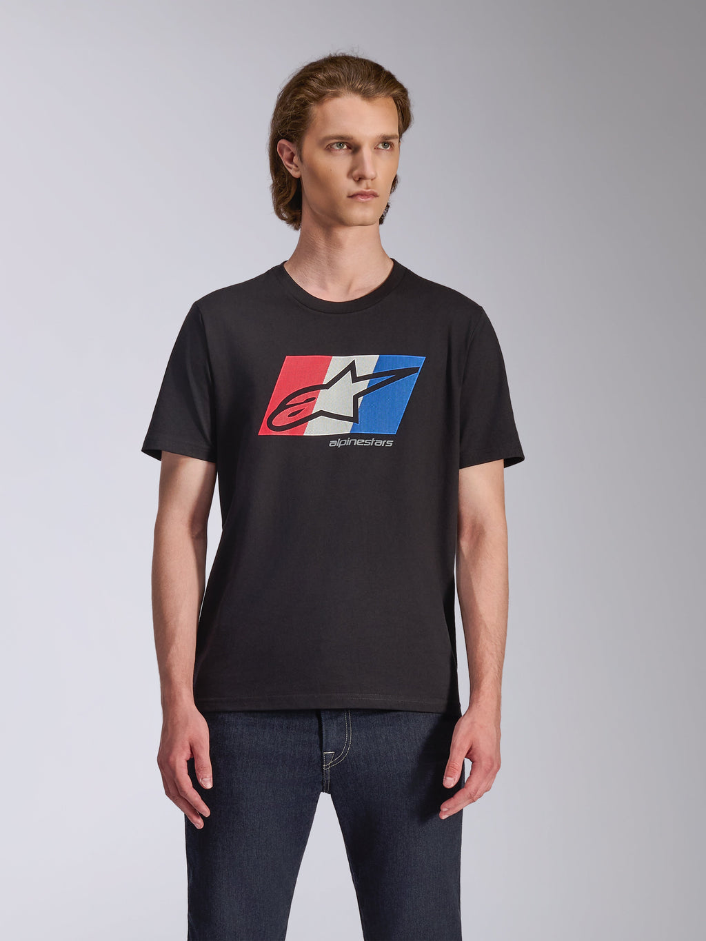 Alpinestars Global CSF Tee, kortärmad t-shirt, svart, röd, vit och blå Ageless-logotypgrafik, bärs av manlig modell med mörka jeans, avslappnad motorsport-streetwear