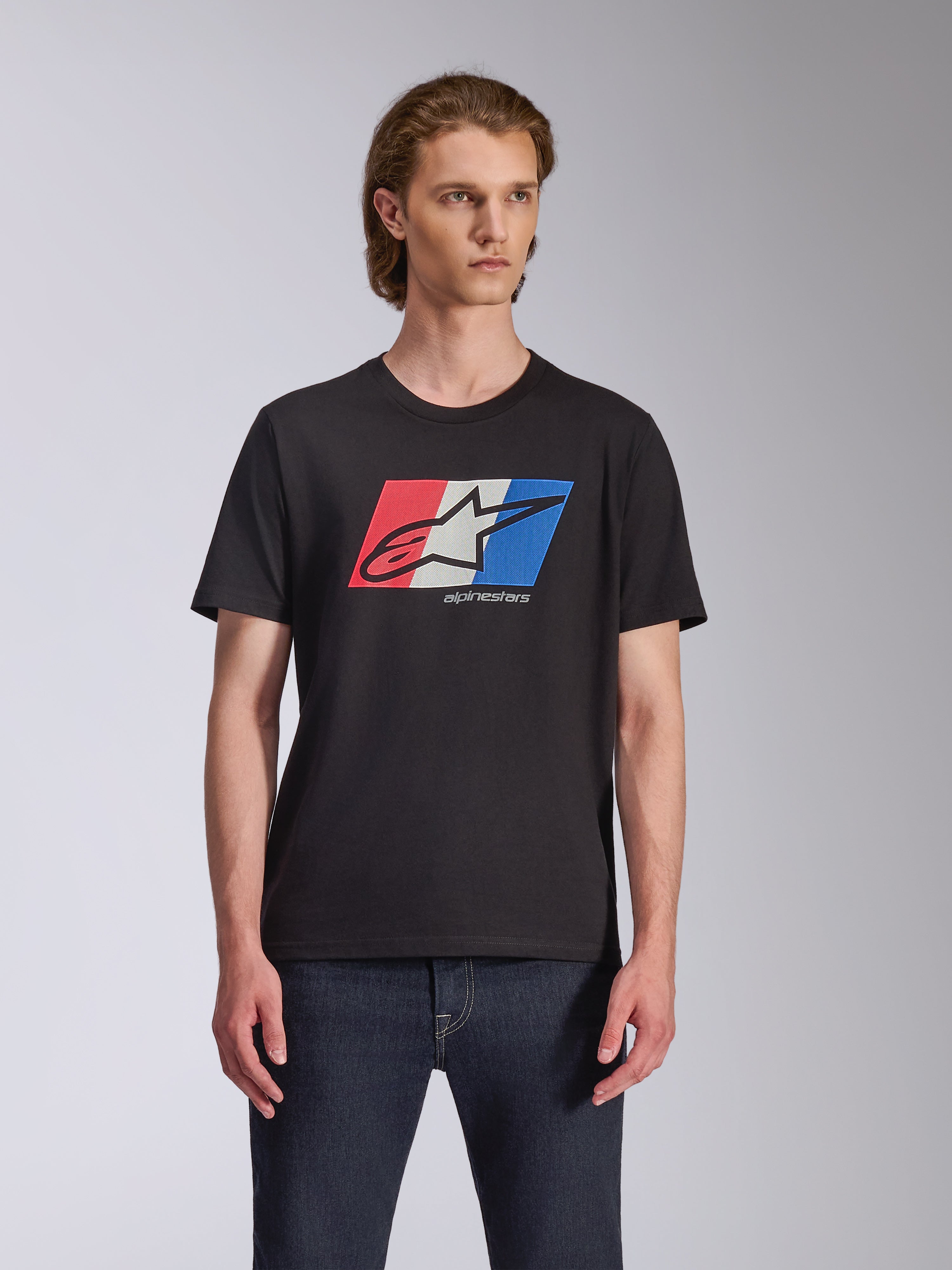 Alpinestars Global CSF Tee, kortärmad t-shirt, svart, röd, vit och blå Ageless-logotypgrafik, bärs av manlig modell med mörka jeans, avslappnad motorsport-streetwear