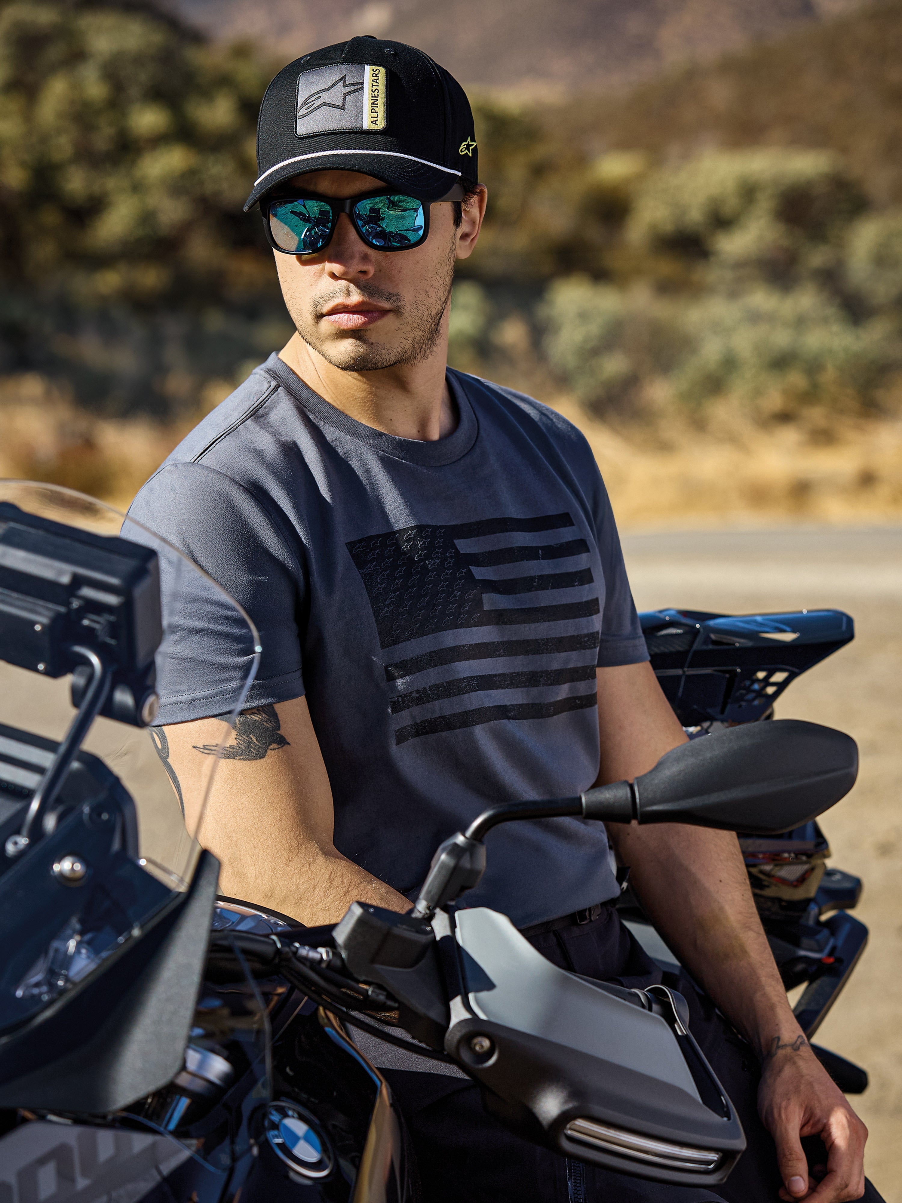 Alpinestars Waving CSF Tee, kortärmad t-shirt, charcoal, med svart grafik av en viftande flagga på bröstet, buren av en man på en motorcykel i solig utomhusmiljö, avslappnad motorsport-livsstil