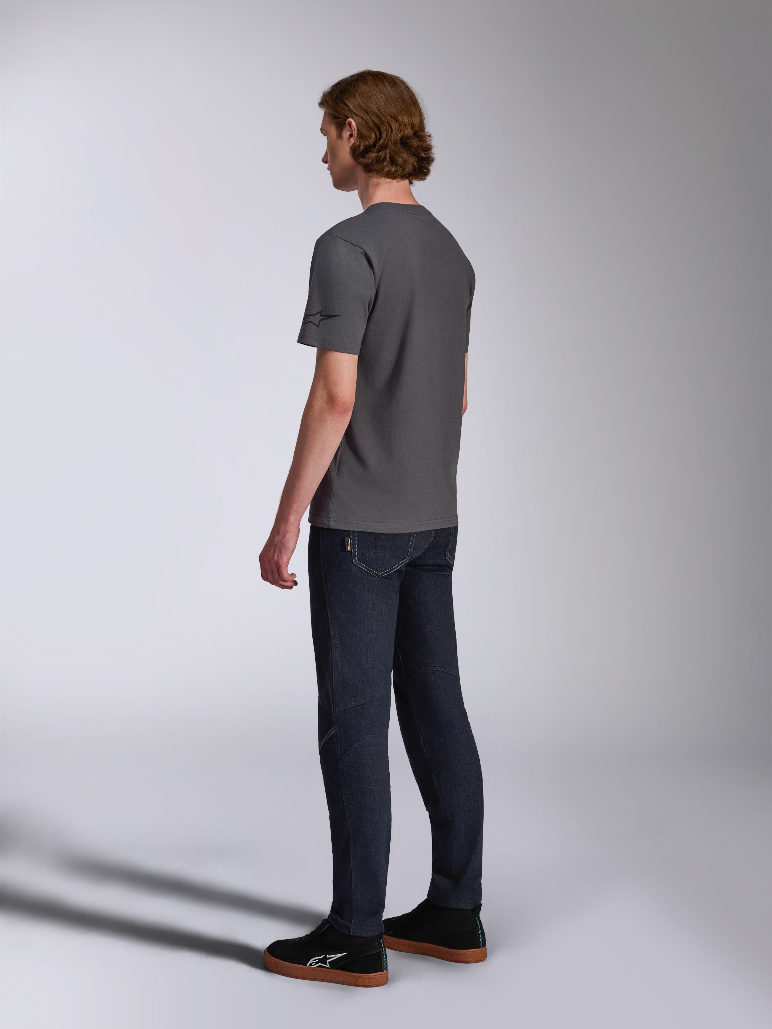 Alpinestars Waving CSF Tee, kortärmad t-shirt, charcoal, sedd bakifrån på modell, liten svart stjärnlogotyp på ärmen, bärs med mörka denimjeans och svarta sneakers, avslappnad motorsport-streetwear