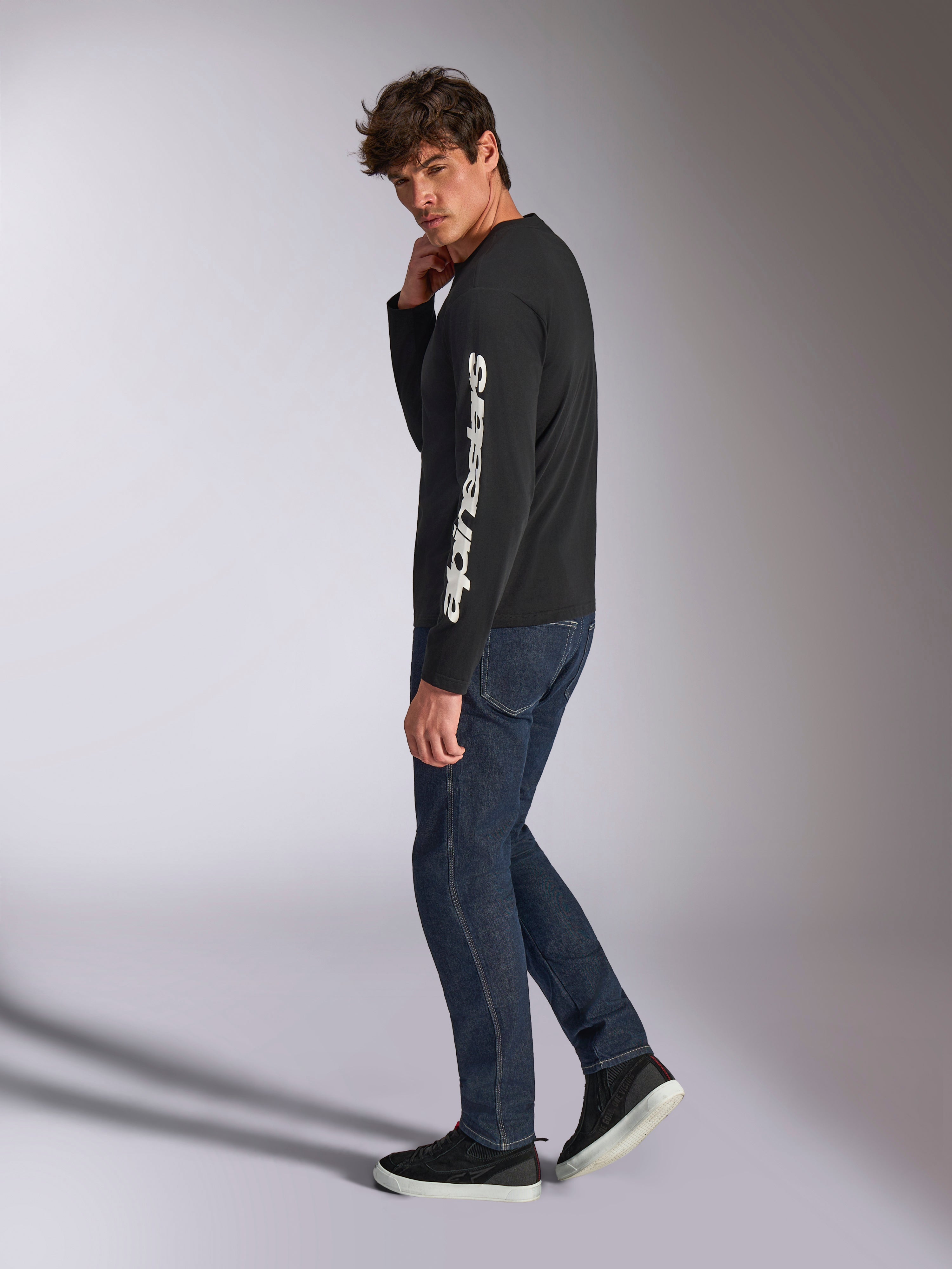 Alpinestars Trackside CSF långärmad t-shirt, svart med vit vertikal Alpinestars-logotyp på ärmen, bärs av en modell med mörk denim och sneakers, avslappnade motorsport-inspirerade sportkläder