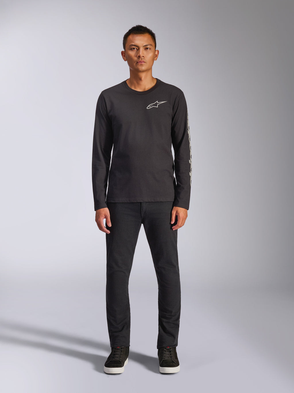 Alpinestars Trackside CSF Tee, långärmad t-shirt, svart, vit Alpinestars-logotyp på bröstet och grafiskt tryck på ärmen, bärs med svarta byxor och sneakers, avslappnad motorsportinspirerad streetwear