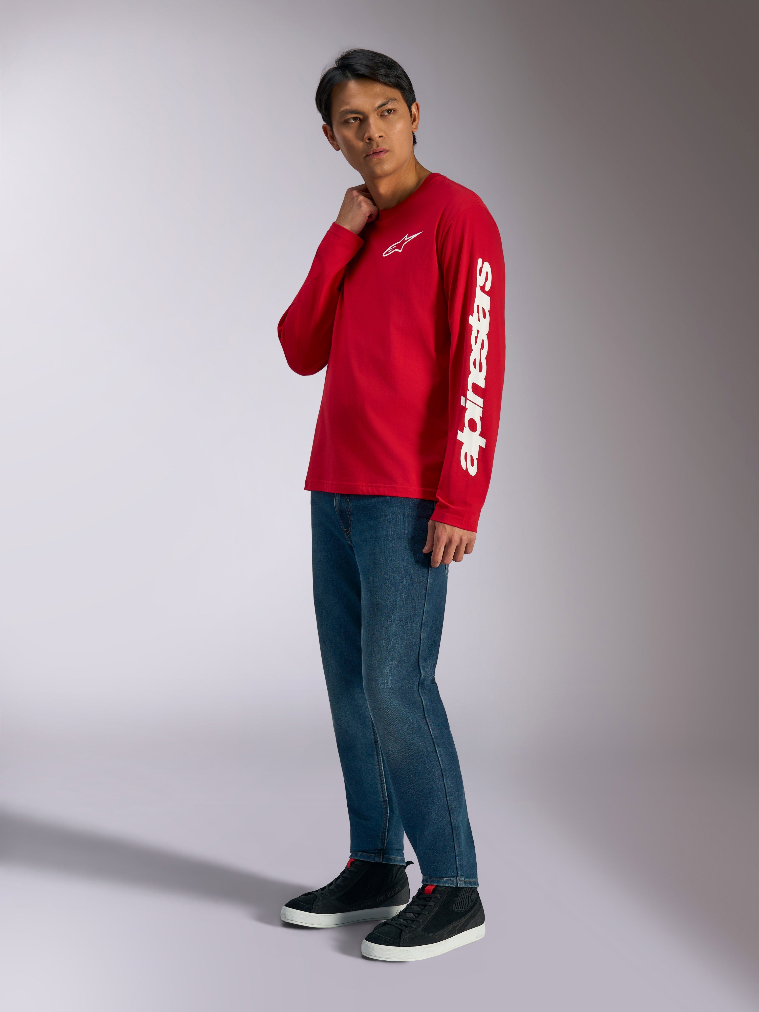 Alpinestars Trackside CSF långärmad t-shirt, röd med vit Alpinestars-logotyp på bröstet och ärmen, bärs av modell med blå jeans och svarta sneakers, avslappnat motorsport-inspirerat streetwear