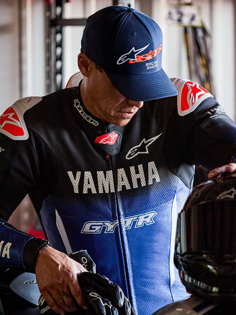 Alpinestars Entitled keps, mörkblå med orange Astars-logotyp och vit 'Racing Ahead'-text, bärs av en förare i ett blått och svart Yamaha-racingskinnställ, baseballkeps med böjd skärm och strukturerad krona