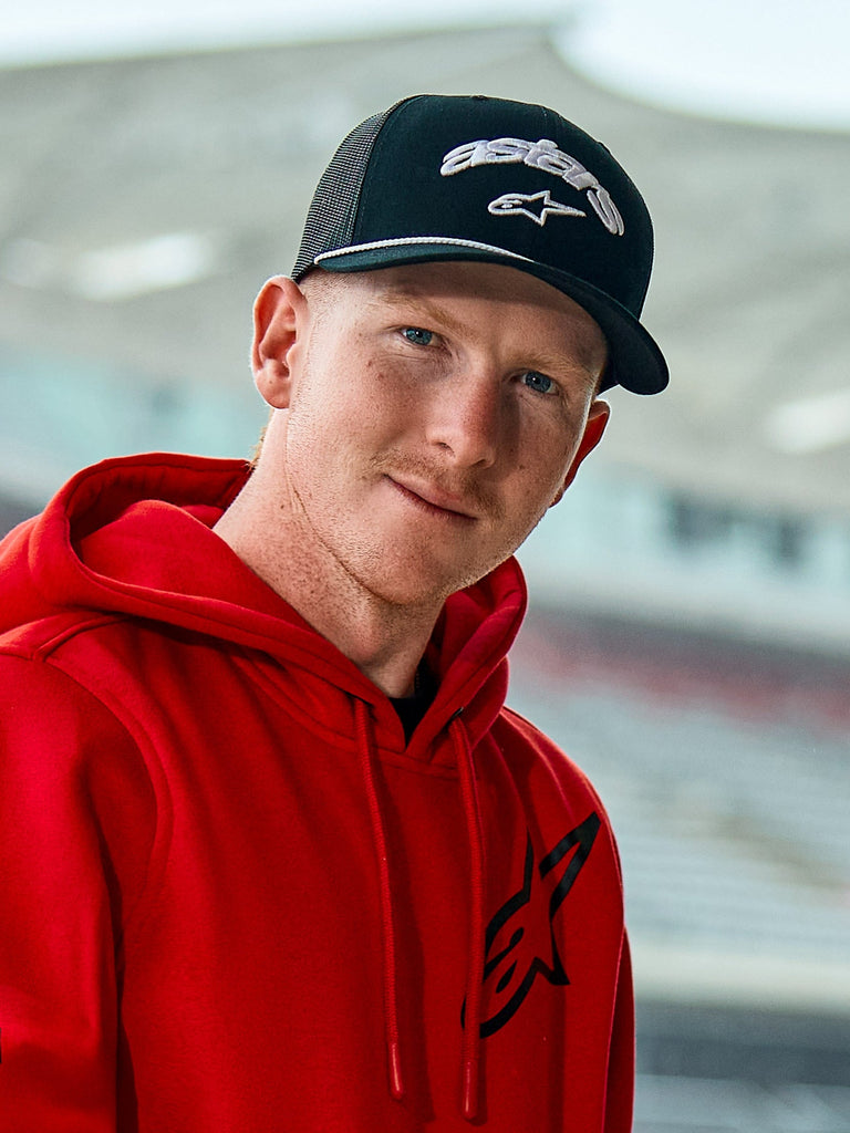 Man i röd Alpinestars-hoodie och Agree Trucker-keps i svart, med vitbroderade Astars-logotyper, vit flätad repdetalj på skärmen och en ventilerande mesh-baksida, vardaglig livsstilsmiljö