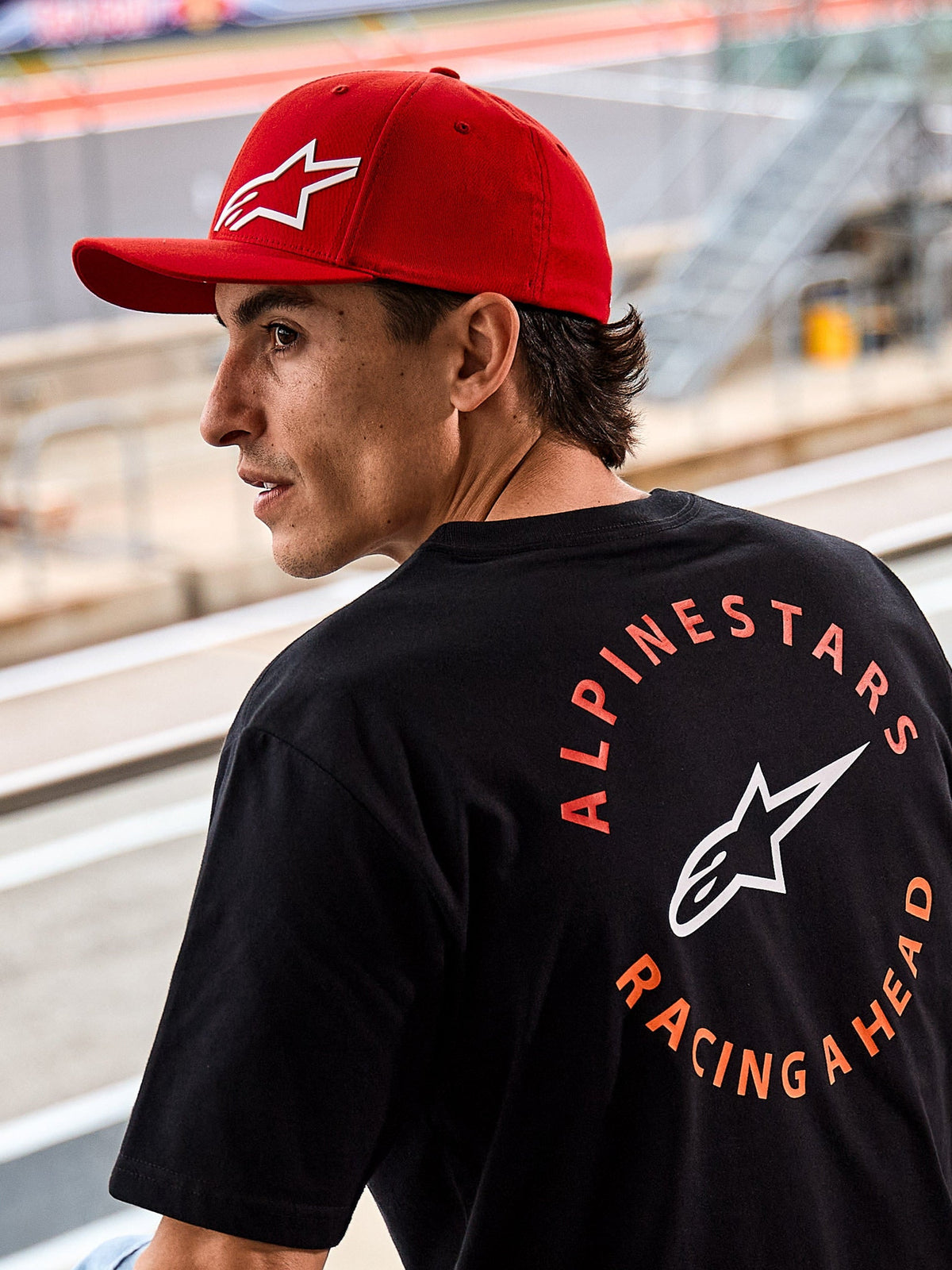 Alpinestars Corp Shift Multi Hat, röd keps med böjd skärm och vit 3D Astars-logotyp, bärs med en svart Alpinestars t-shirt med 'Racing Ahead'-grafik, livsstilsbild i motorsportmiljö vid en racingbana