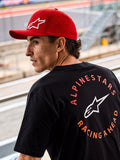 Alpinestars Corp Shift Multi Hat, röd keps med böjd skärm och vit 3D Astars-logotyp, bärs med en svart Alpinestars t-shirt med 'Racing Ahead'-grafik, livsstilsbild i motorsportmiljö vid en racingbana