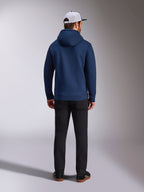 Alpinestars Frontal Hoodie, marinblå, ryggvy av man som bär en avslappnad pullover-fleecehuvtröja, med ribbade muddar och fåll, matchad med svarta byxor och en vit meshkeps, motorsportinspirerad streetwear