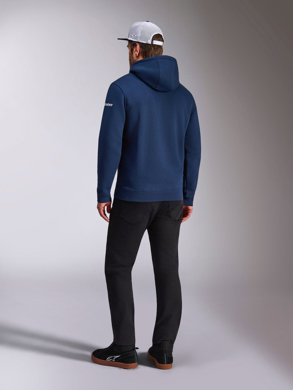 Alpinestars Frontal Hoodie, vy bakifrån av en marinblå pullover-fleecehuvtröja, vit ordlogotyp på ärmen, matchad med svarta byxor och en vit trucker-keps i mesh, avslappnad motorsport-inspirerad streetwear