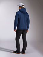 Alpinestars Frontal Hoodie, vy bakifrån av en marinblå pullover-fleecehuvtröja, vit ordlogotyp på ärmen, matchad med svarta byxor och en vit trucker-keps i mesh, avslappnad motorsport-inspirerad streetwear