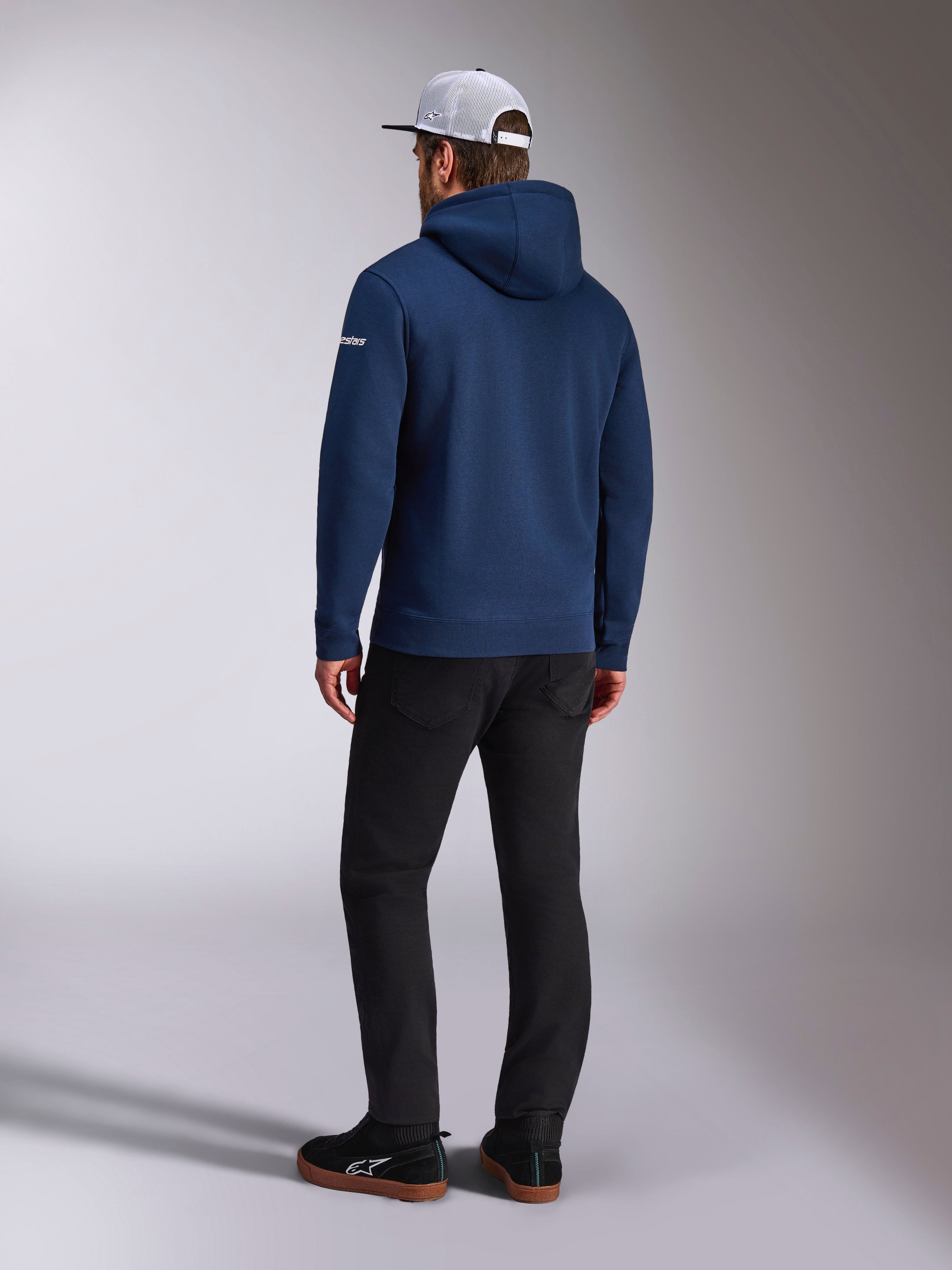 Alpinestars Frontal Hoodie, vy bakifrån av en marinblå pullover-fleecehuvtröja, vit ordlogotyp på ärmen, matchad med svarta byxor och en vit trucker-keps i mesh, avslappnad motorsport-inspirerad streetwear