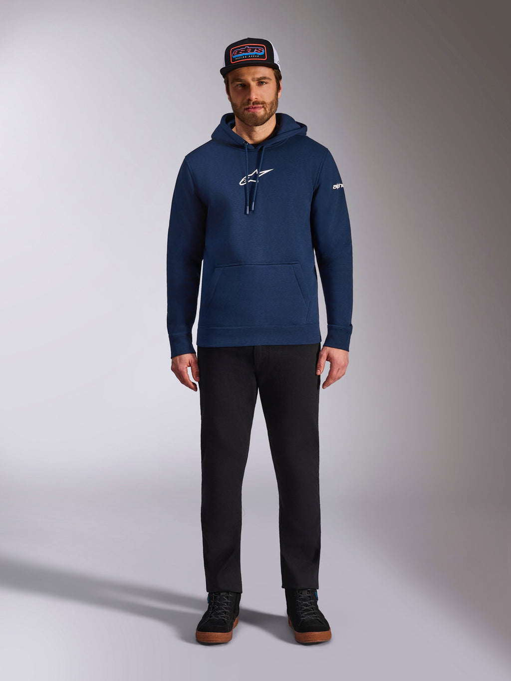 Manlig modell bär Alpinestars Frontal Hoodie i Navy, en fleece-huvtröja med vit Astars-logotyp på bröstet och ordlogotyp på ärmen, utrustad med känguruficka och huva med dragsko, matchat med svarta byxor och en keps med logotyp, avslappnat motorsport-streetwear