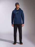 Manlig modell bär Alpinestars Frontal Hoodie i Navy, en fleece-huvtröja med vit Astars-logotyp på bröstet och ordlogotyp på ärmen, utrustad med känguruficka och huva med dragsko, matchat med svarta byxor och en keps med logotyp, avslappnat motorsport-streetwear