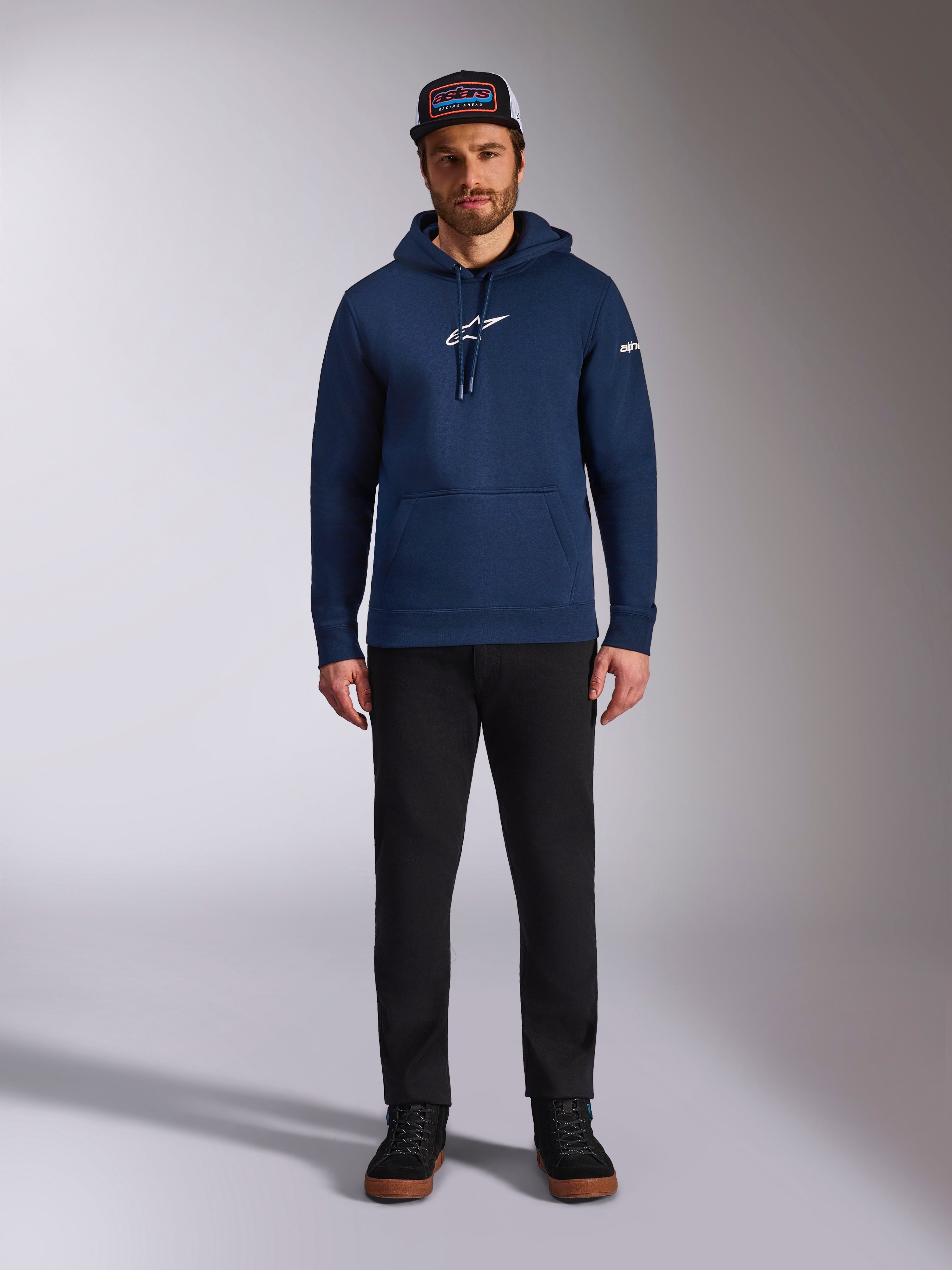 Manlig modell bär Alpinestars Frontal Hoodie i Navy, en fleece-huvtröja med vit Astars-logotyp på bröstet och ordlogotyp på ärmen, utrustad med känguruficka och huva med dragsko, matchat med svarta byxor och en keps med logotyp, avslappnat motorsport-streetwear