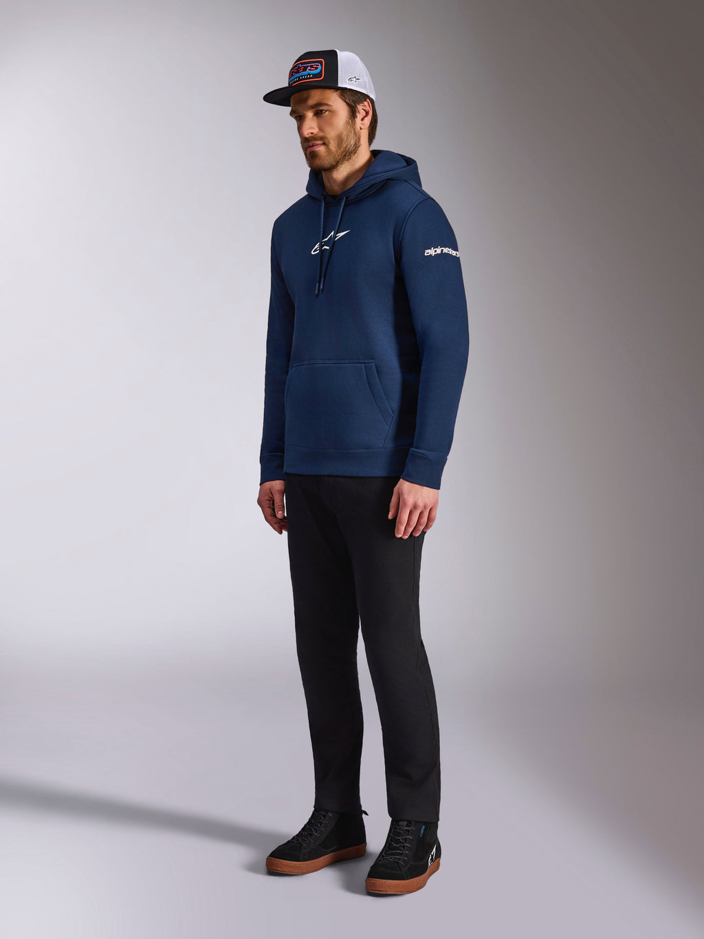 Alpinestars Frontal Hoodie i marinblått, avslappnad huvtröja i fleece med vit Astars-logotyp på bröstet och ordlogotyp på ärmen, stylad med truckerkeps och svarta byxor, moderna motorsport-inspirerade livsstilskläder.