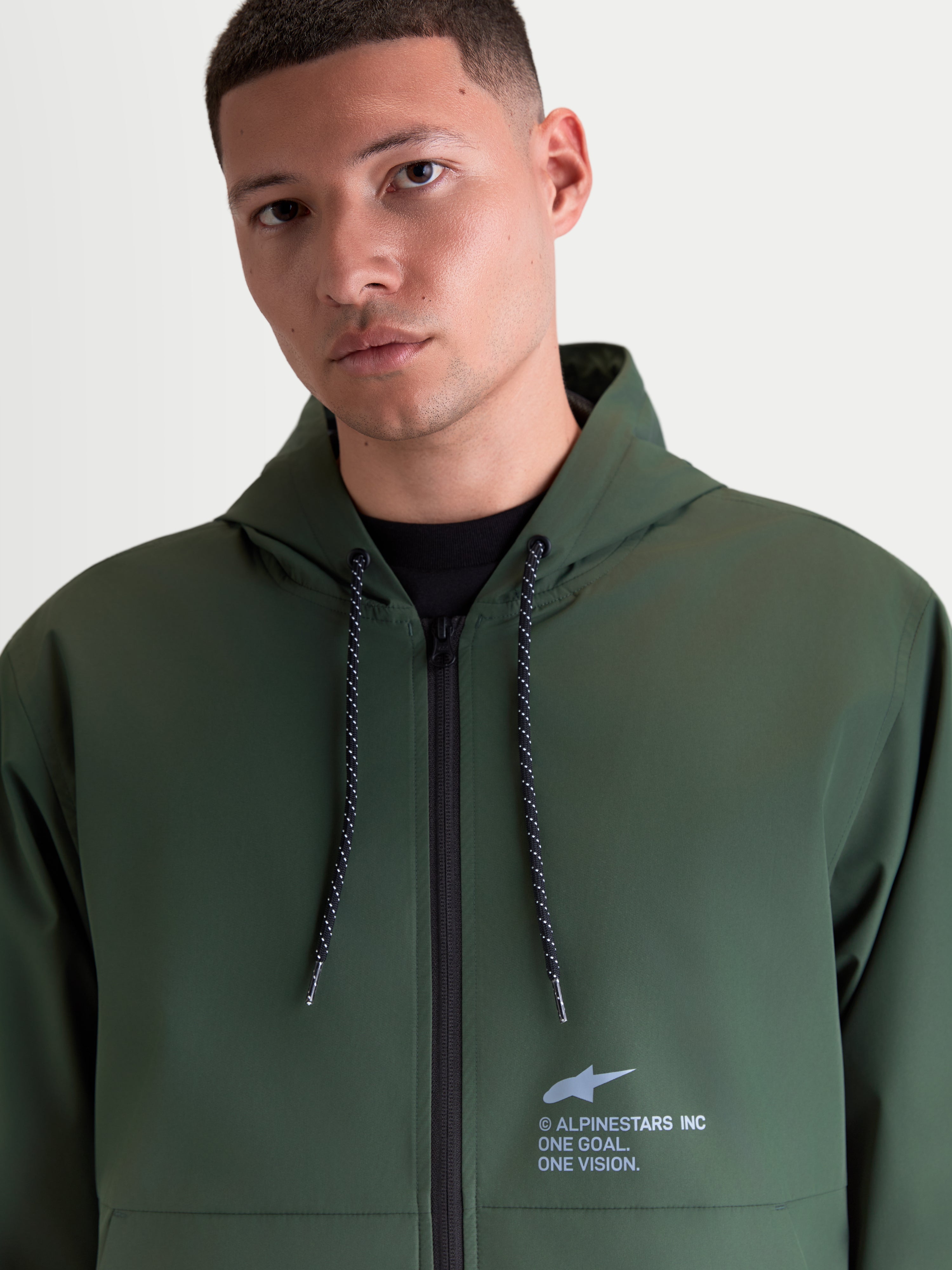 Alpinestars Speeder Resolve vindstopp jacka, lätt grön sportjacka för herrar med huva, spräckliga dragskor och signaturbranding på bröstet, buren av en manlig modell.