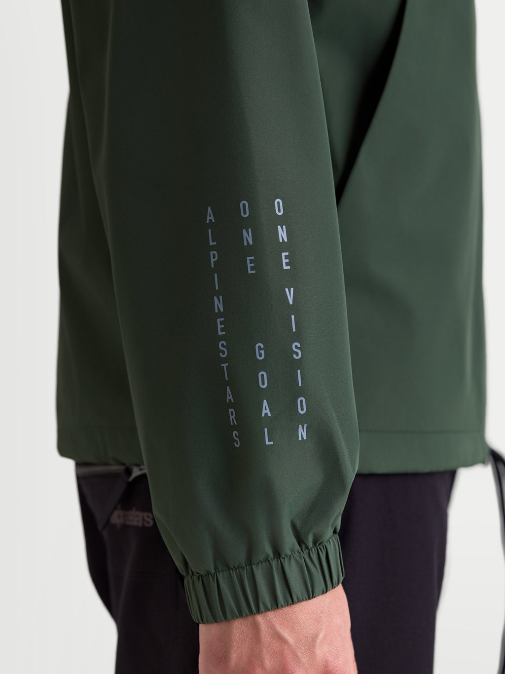 Alpinestars Speeder Resolve vindstoppjacka, grön lättviktsjacka i sportswear-stil, närbild på ärm med vertikal Alpinestars-branding och mottot One Goal One Vision, elastisk mudd vid ärmslut.