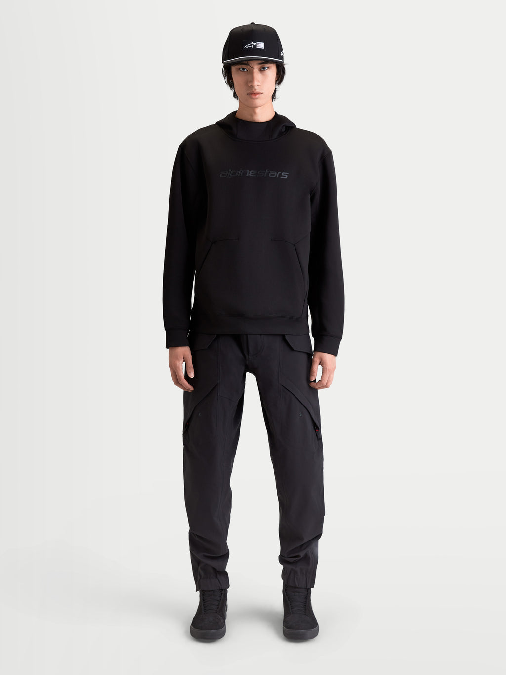 Alpinestars Datrix huvtröja, premium sporttröja för herrar, svart, med tonalt Alpinestars HD-tryck, dubbelvävd tri-blend-fleece och ficka med dartdetalj, visas på modell för en motorsportinspirerad vardagsstil.