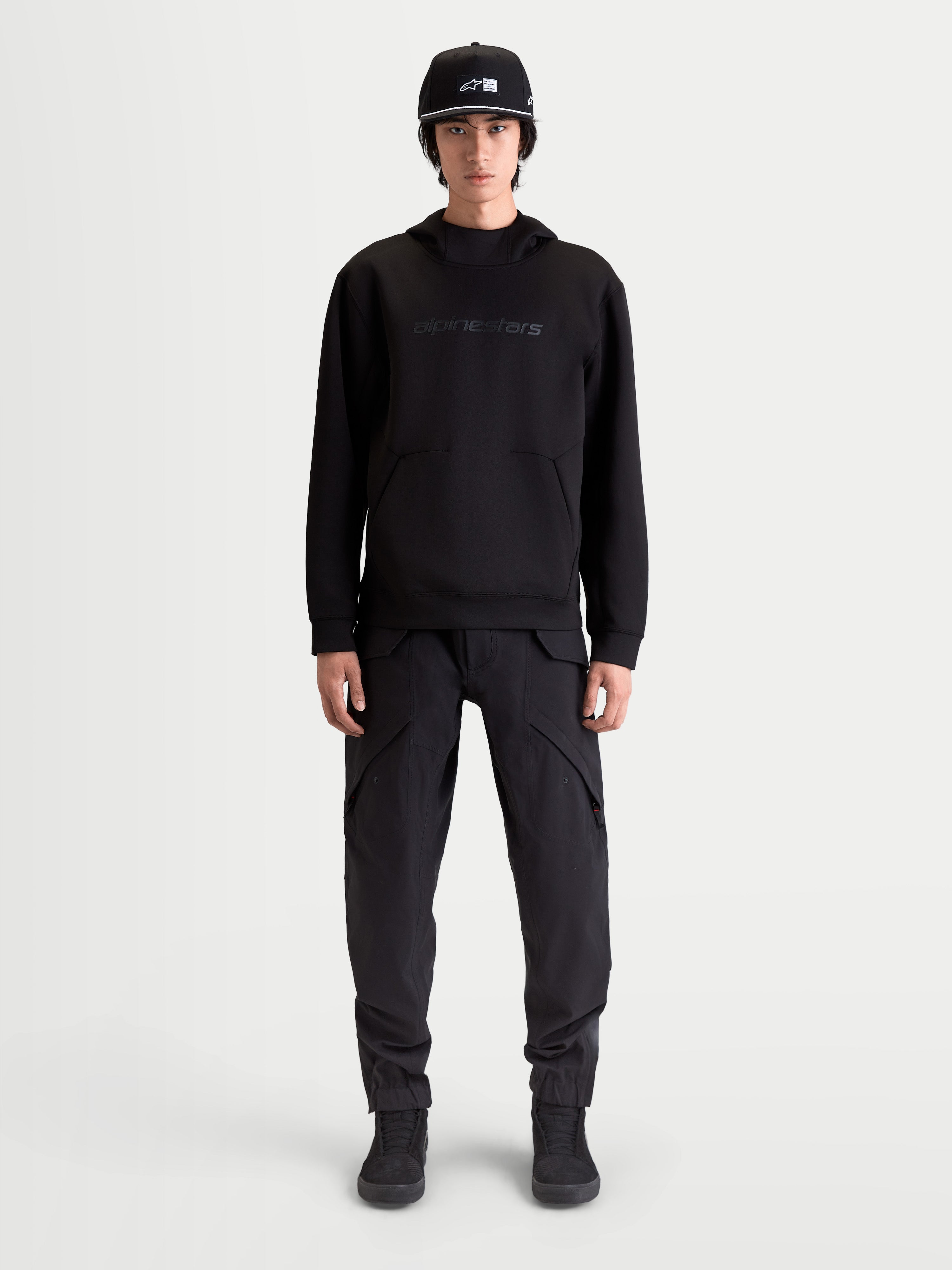 Alpinestars Datrix huvtröja, premium sporttröja för herrar, svart, med tonalt Alpinestars HD-tryck, dubbelvävd tri-blend-fleece och ficka med dartdetalj, visas på modell för en motorsportinspirerad vardagsstil.