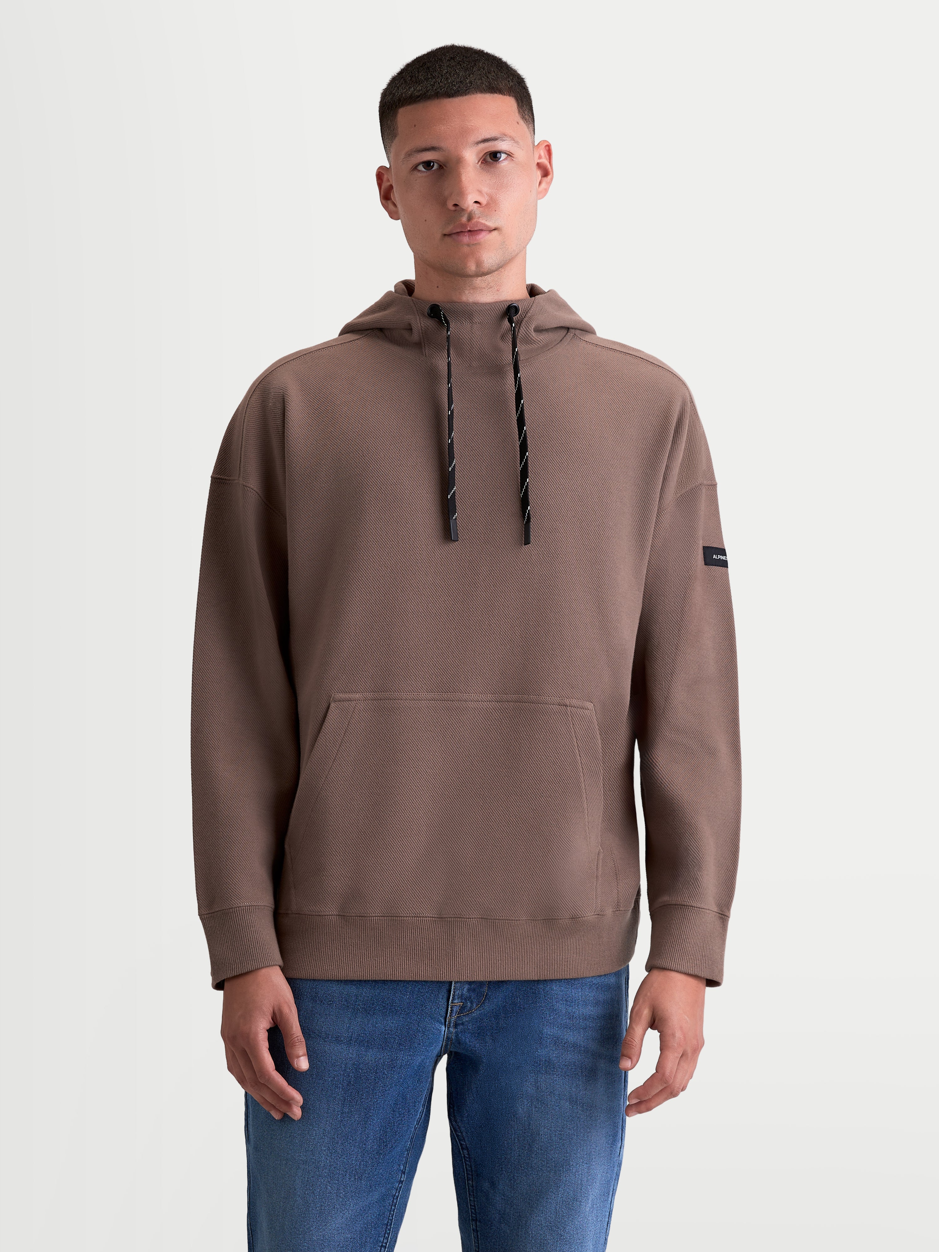 Alpinestars Camino Textured Oversized Hoodie, Men's Sportswear Pullover, Taupe Brown, featuring a high collar, kangaroo pocket, and textured knit twill fleece for a modern motorsport-inspired look. On-model photo showing the relaxed silhouette with two-tone drawcords and a subtle sleeve logo.