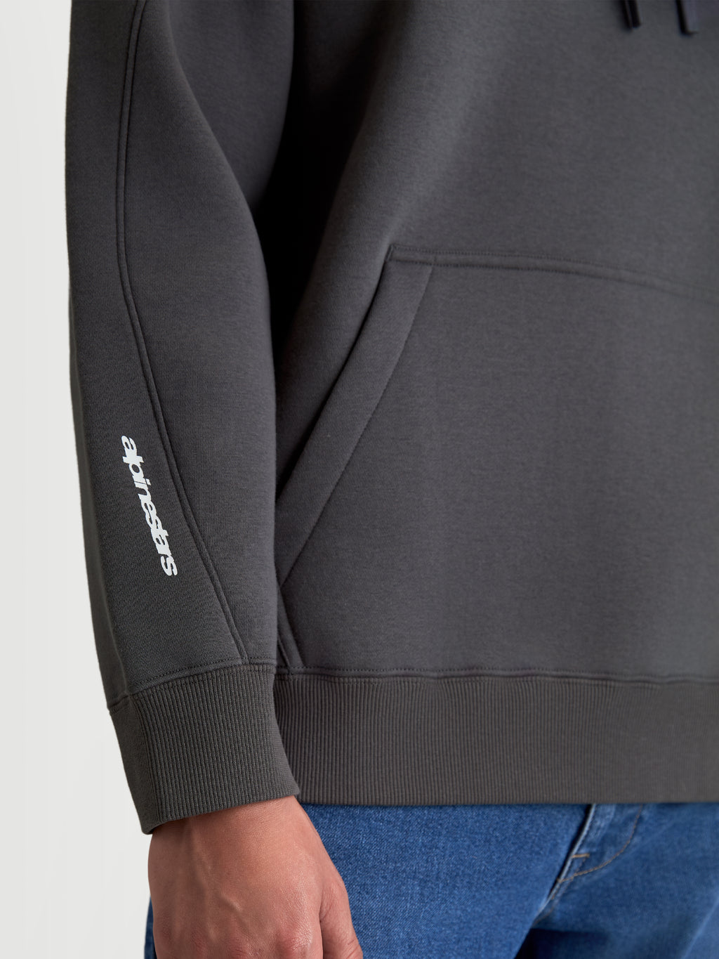 Alpinestars Prelude Oversized huvtröja, mörkgrå sportswear fleece-pullover, detaljvy som visar ärmsömmar, ribbad mudd och vit Alpinestars-logotyp på armen, avslappnad passform med känguruficka.
