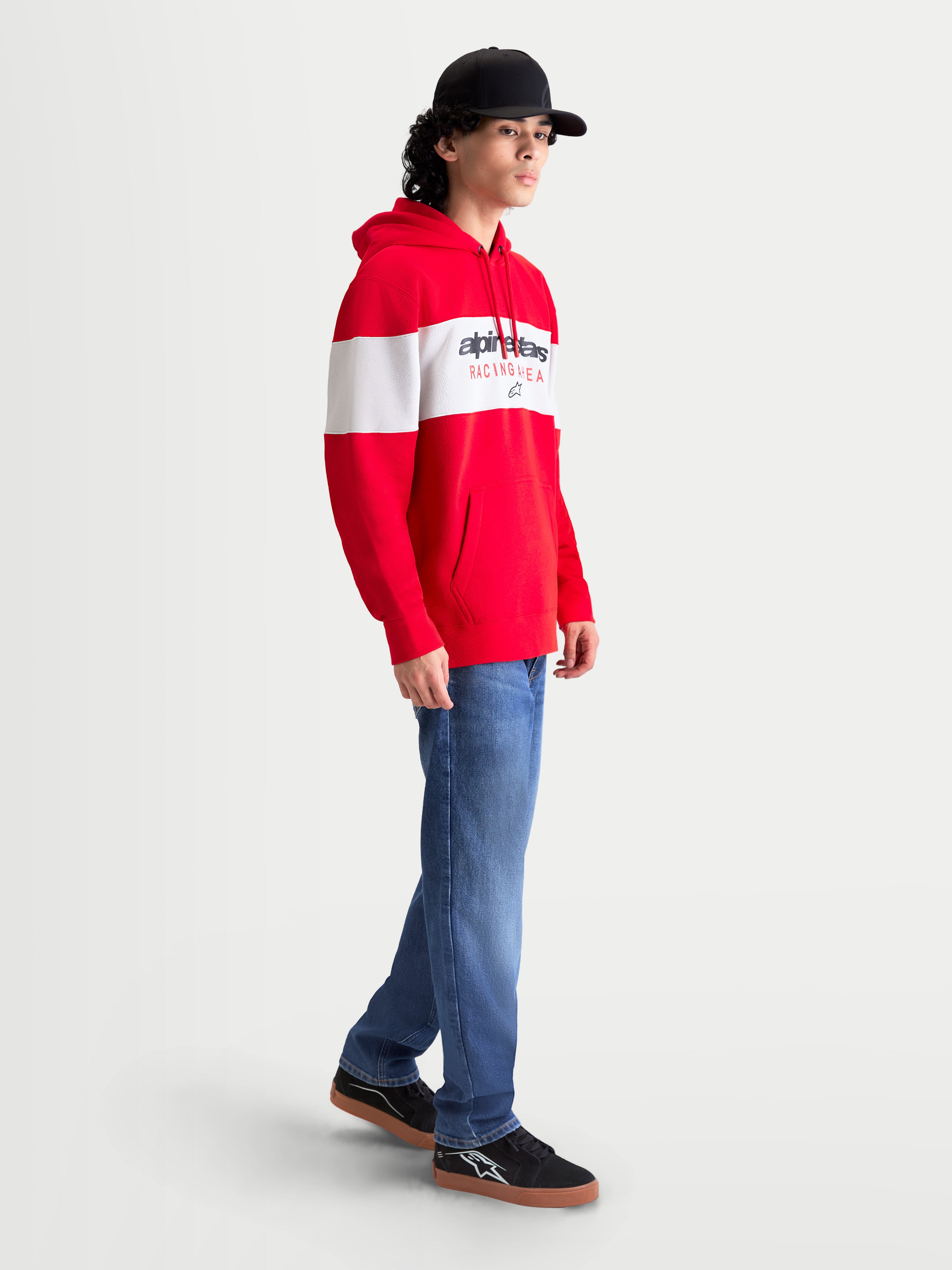Alpinestars Cubic huvtröja, röd och vit herr-huvtröja i fleece med färgblockerad design och Alpinestars ordlogotyp på bröstet. Sidovy på modell som visar huva med dragsnöre, känguruficka och ribbade muddar för en avslappnad stil utanför banan.