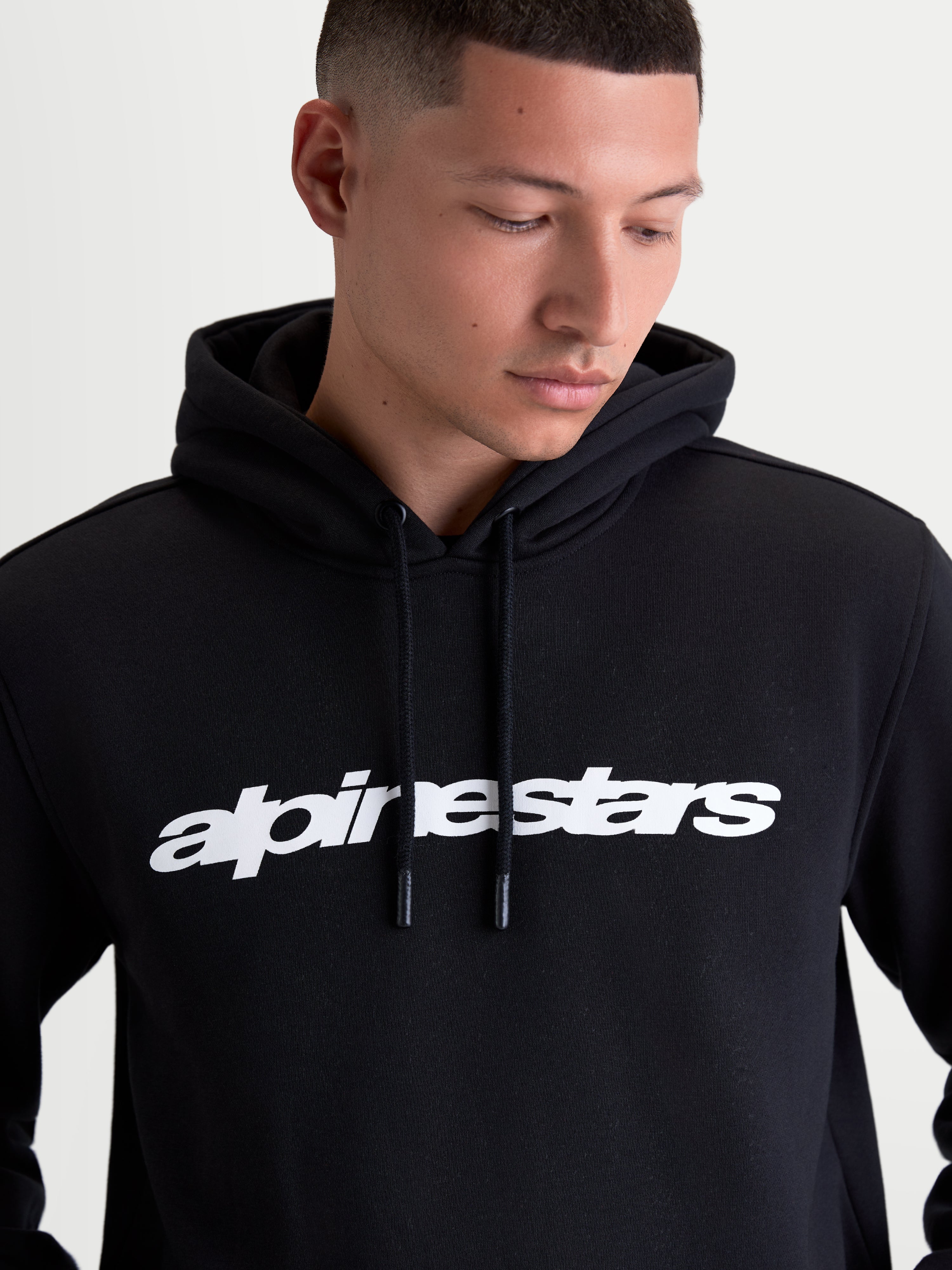 Alpinestars Race Wordmark huvtröja, svart och vit, buren av en modell, sportig pulloverfleece med tydlig vit logotyp på bröstet, självfodrad huva och dragsko med doppade ändar, bomull och återvunnen polyester.