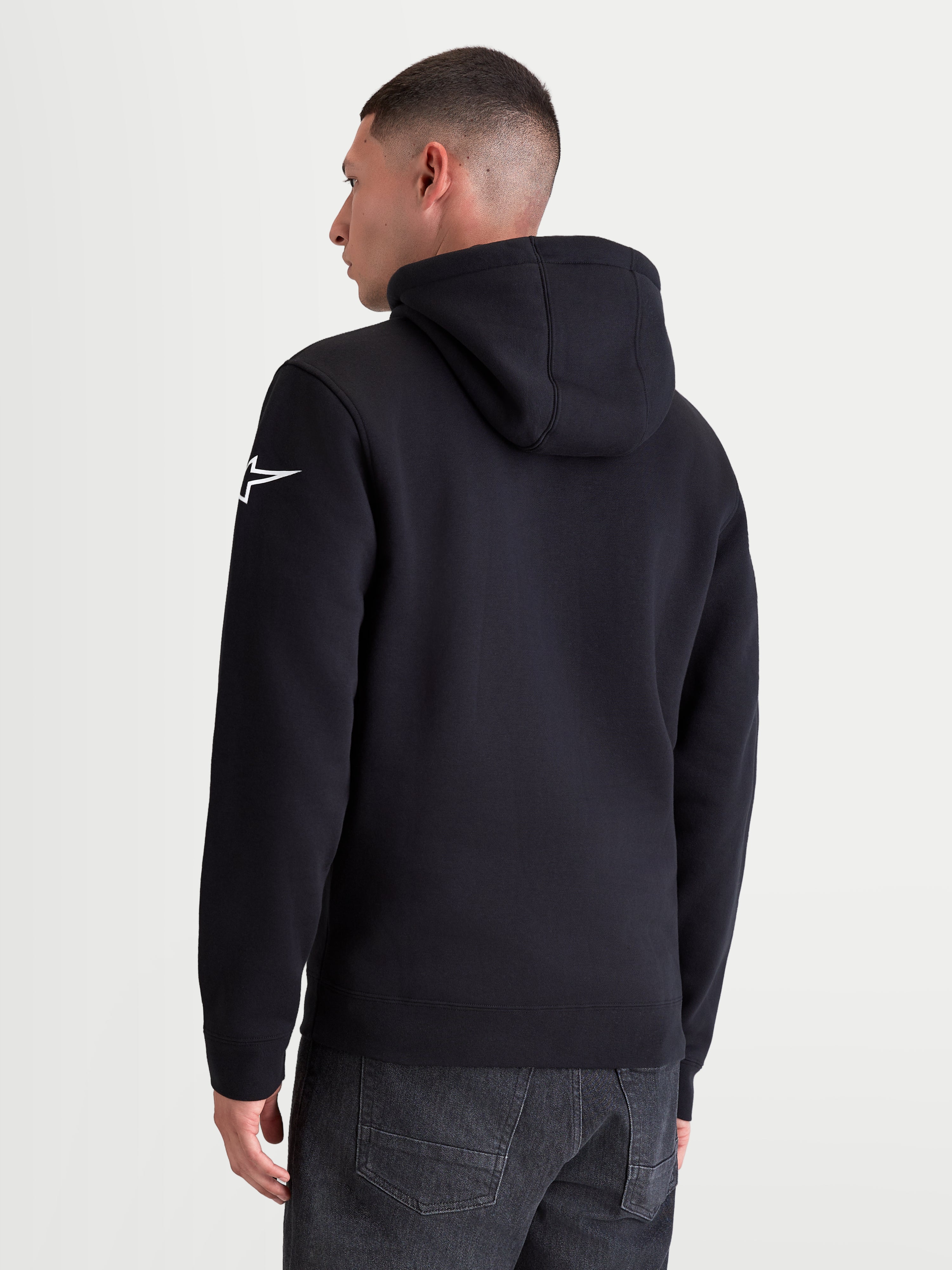 Alpinestars Race Wordmark huvtröja, sportig pullover i fleece, ryggvy på modell, svart och vit, med vit Ageless-stjärnlogotyp på vänster ärm, fodrad huva och ribbade muddar. Tillverkad av bomull och återvunnen polyester för bekväm vardagsanvändning.