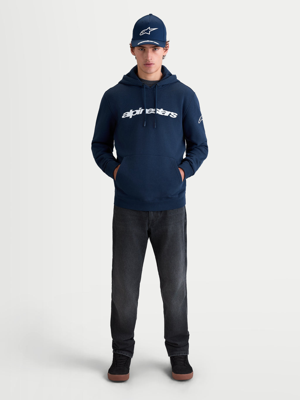 Alpinestars Race Wordmark huvtröja, sportig huvtröja i fleece, marinblå och vit, stor vit wordmark-logotyp på bröstet och Ageless-logotyp på ärmen, livsstilsbild på modell för en avslappnad motorsport-inspirerad look.