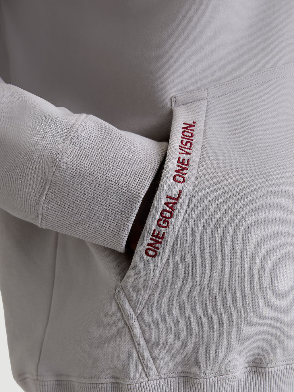 Alpinestars Pioneer Oversized huvtröja, cementgrå och röd, närbild på känguruficka med broderad text 'ONE GOAL. ONE VISION.' och ribbad mudd, premium sportswear för motorsportsentusiaster