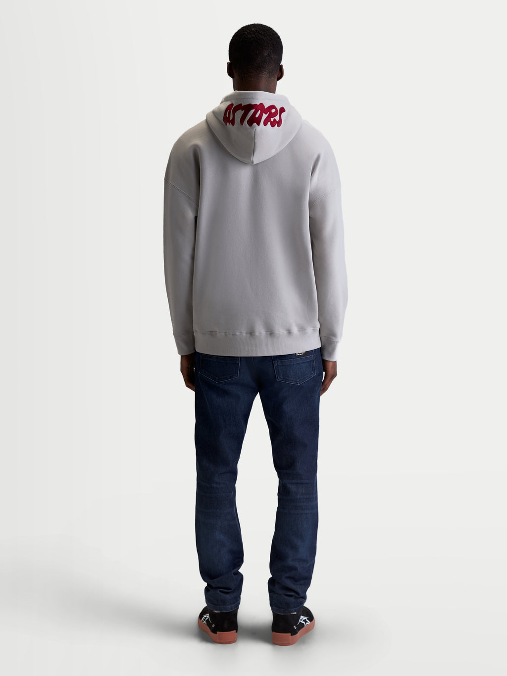 Alpinestars Pioneer Oversized huvtröja, bekväm sportswear-huvtröja i fleece för herr och dam, cementgrå och röd, vy bakifrån på modell som framhäver röd broderad ASTARS-logotyp på huvan och en avslappnad oversizepassform.