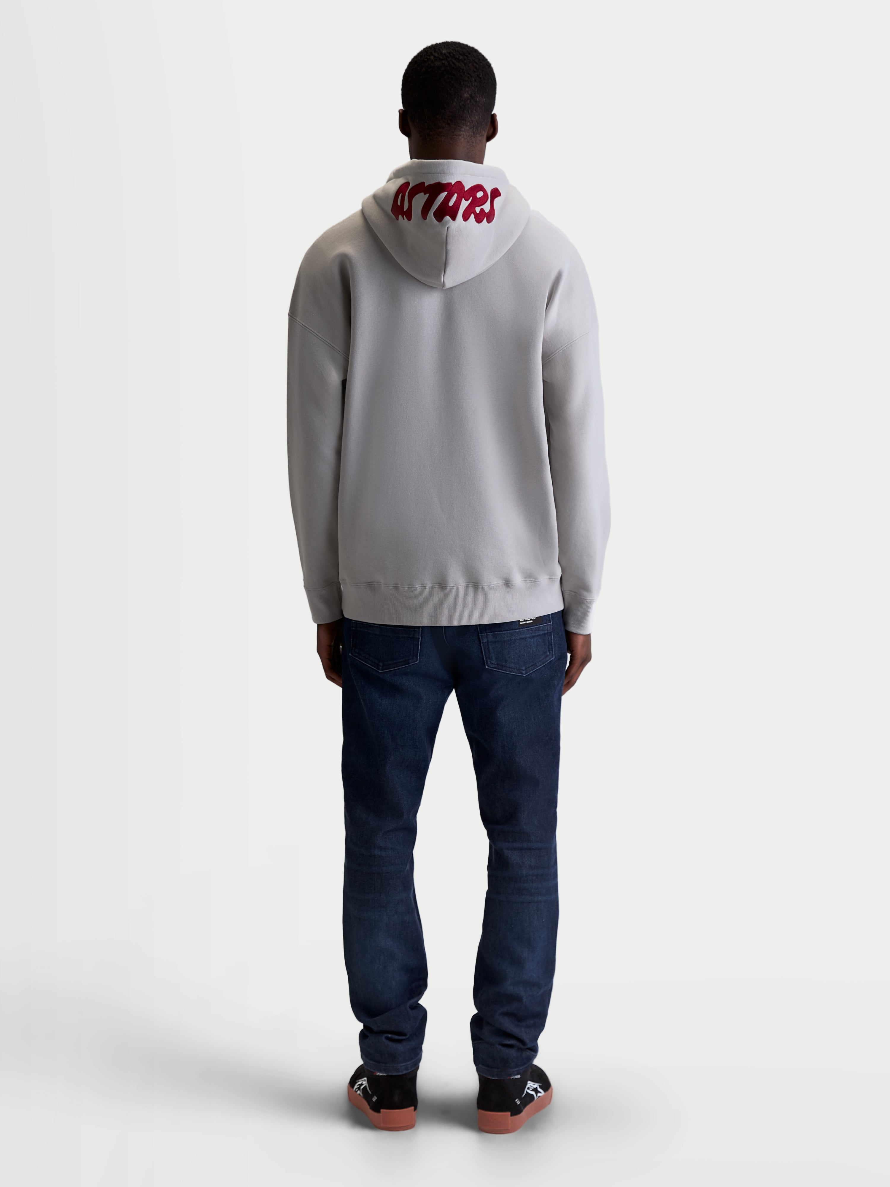 Alpinestars Pioneer Oversized huvtröja, bekväm sportswear-huvtröja i fleece för herr och dam, cementgrå och röd, vy bakifrån på modell som framhäver röd broderad ASTARS-logotyp på huvan och en avslappnad oversizepassform.