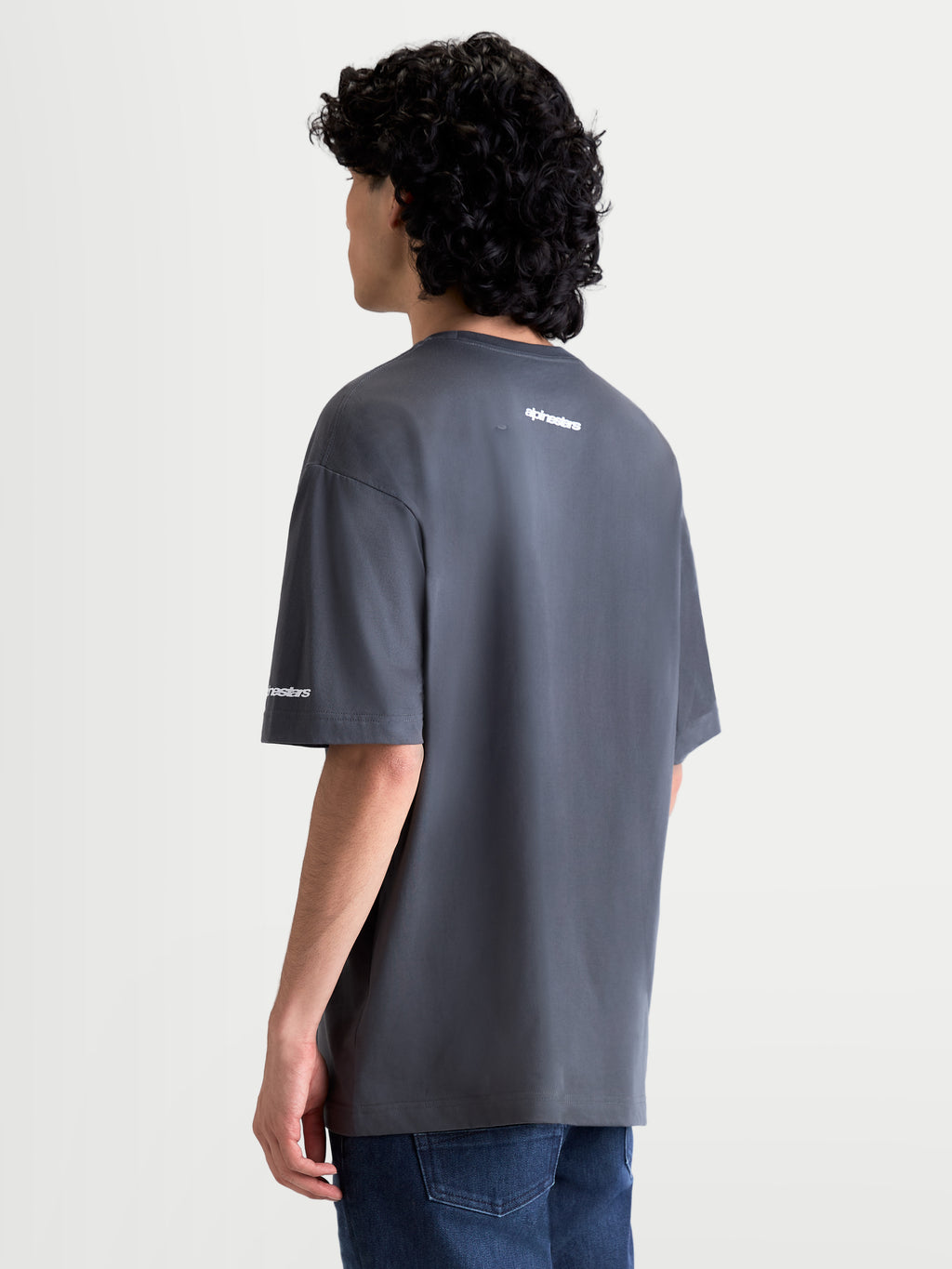 Alpinestars Mandate Oversized CSF t-shirt, kortärmad mörkgrå t-shirt med vit pufftryckt logotyp på ryggen och ärmen, oversized avslappnad passform, bomulls- och polyesterblandning, sedd bakifrån på modell, bekväm motorsport-streetwear.
